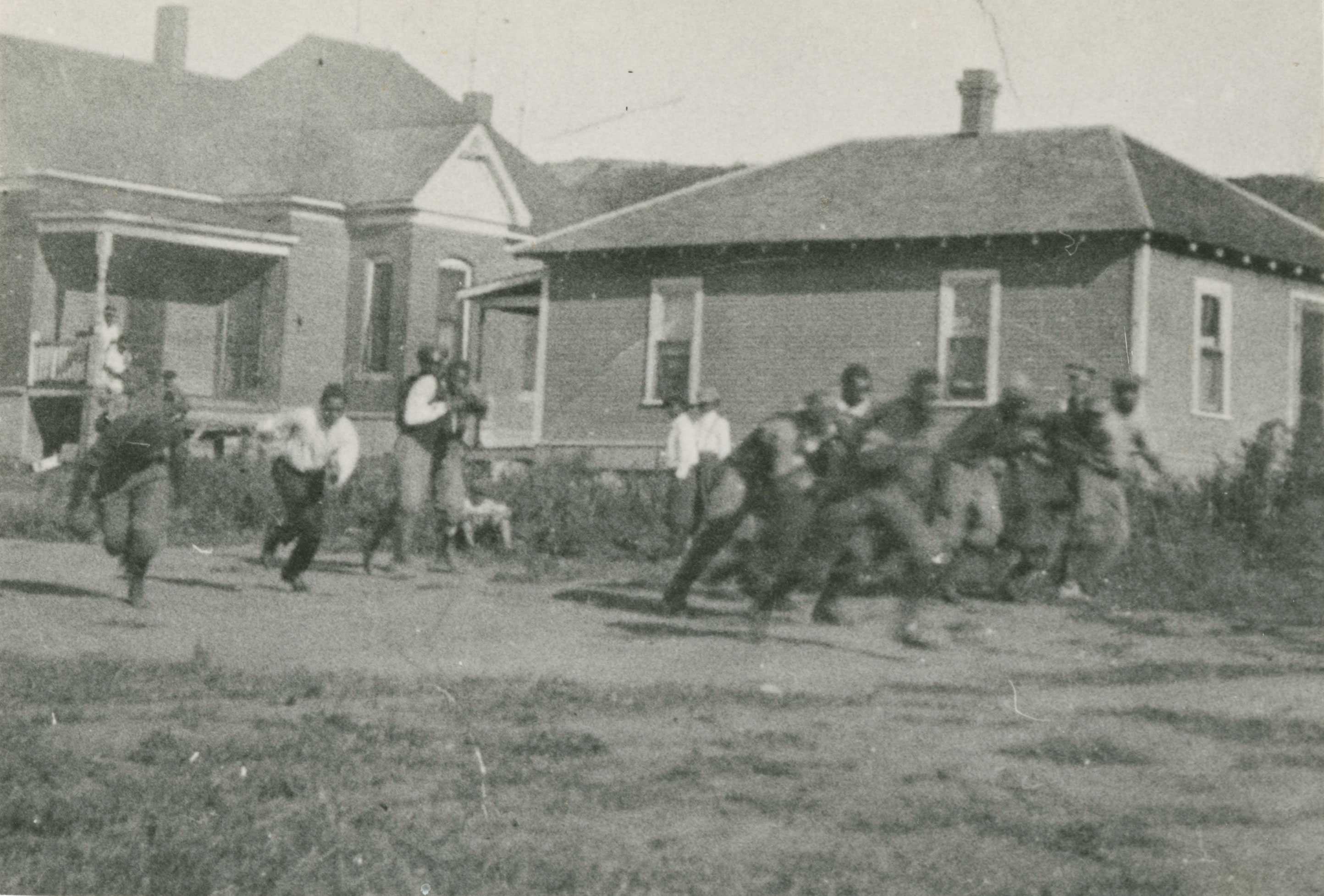 A black-and-white print of children playing football on a dirt lot with others watching from the sidelines and a porch in the background.