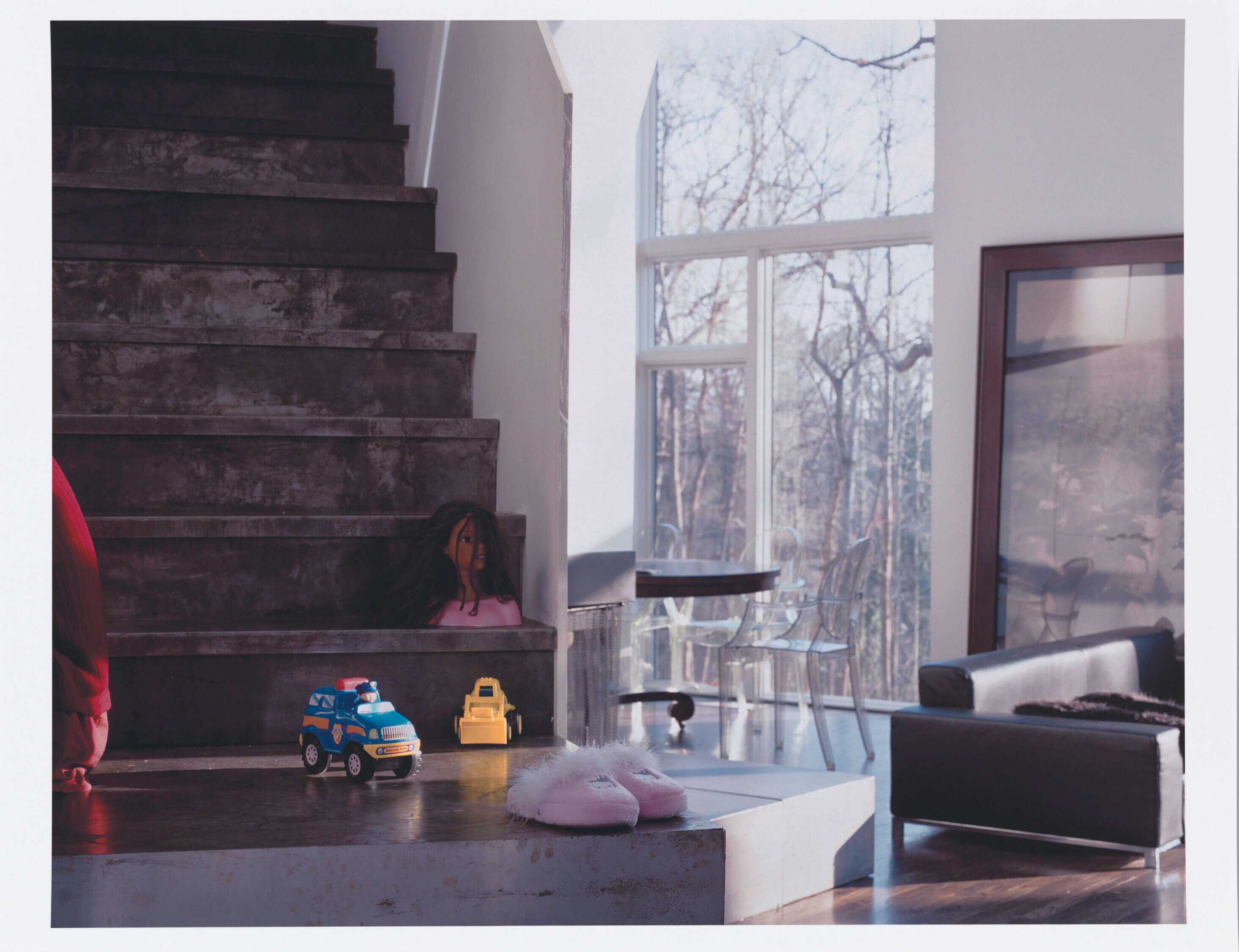 A color photograph of the interior of a modern house. To left is a staircase that leads down to a landing on which there are two (2) child's toy trucks and a pair of pink fuzzy slippers. On the first stair is a doll's head with hair.  To right is the living space of the home, including a leather sofa, a large framed artwork, and a wooden dining table with clear plastic chairs. Behind the table are large pane glass windows with an expanse of leafless trees visible through the windows.