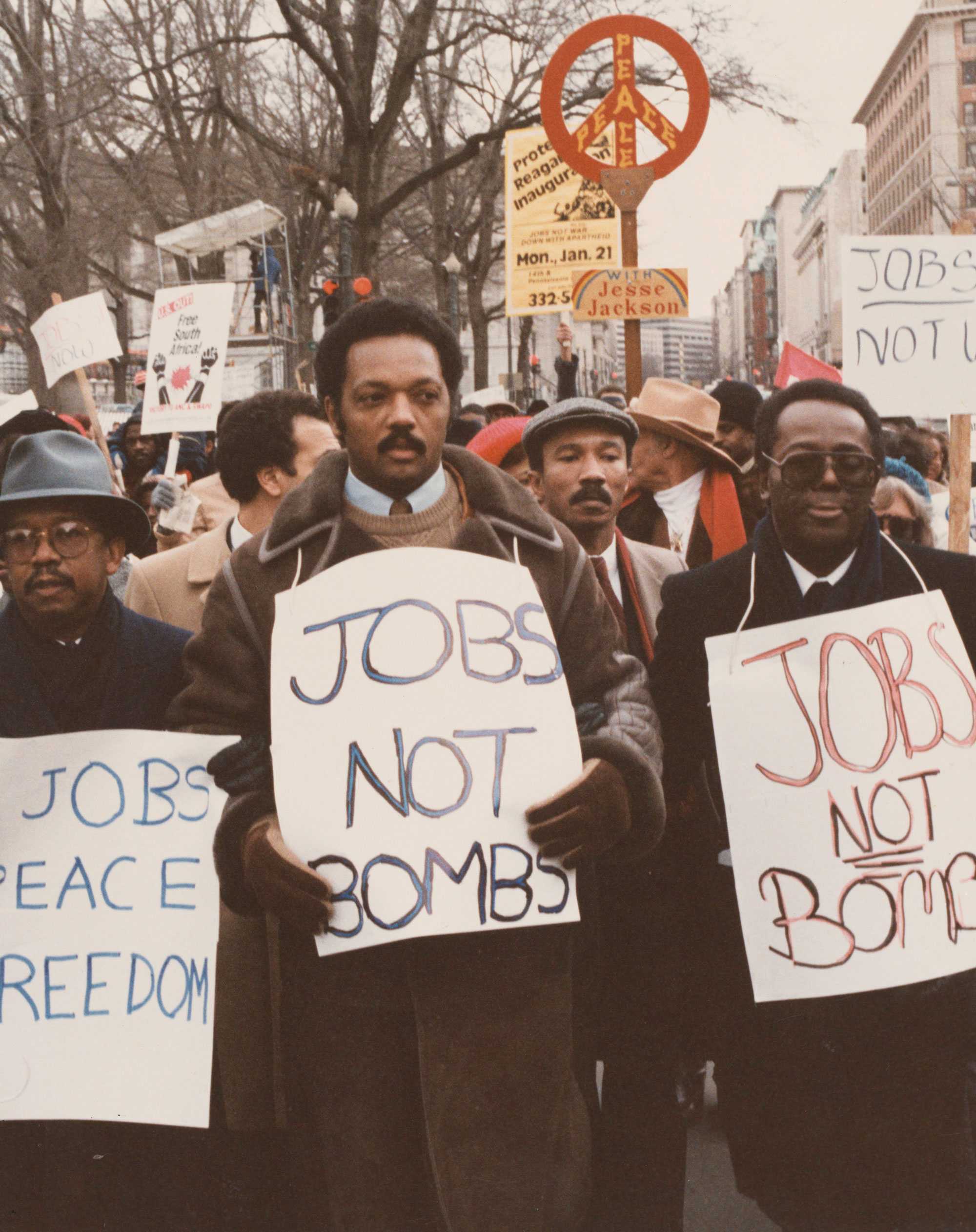 Color photo of Jesse Jackson at a 1975 protest outside the White House, holding a "Jobs not Bombs" sign. Surrounded by supporters with similar signs.