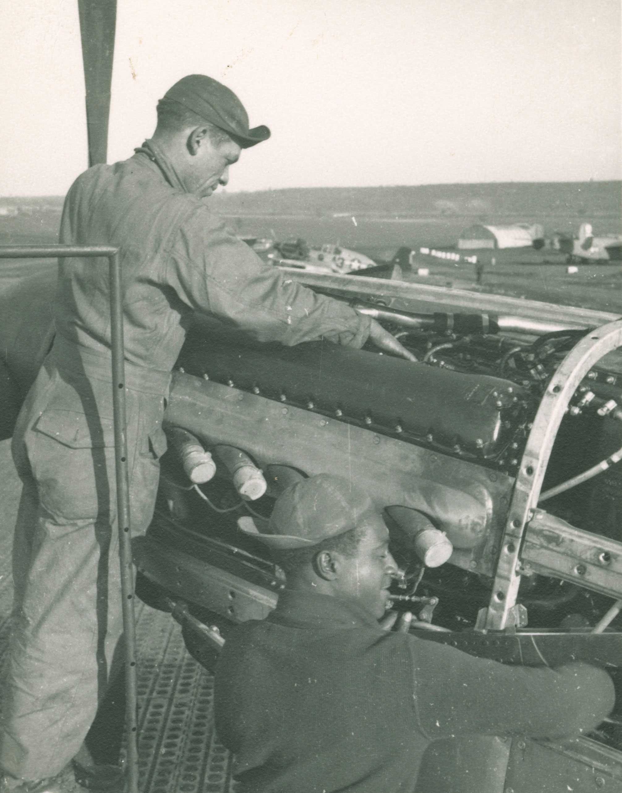 A black-and-white photo of two Tuskegee Airmen servicing an airplane on a pierced steel airstrip.