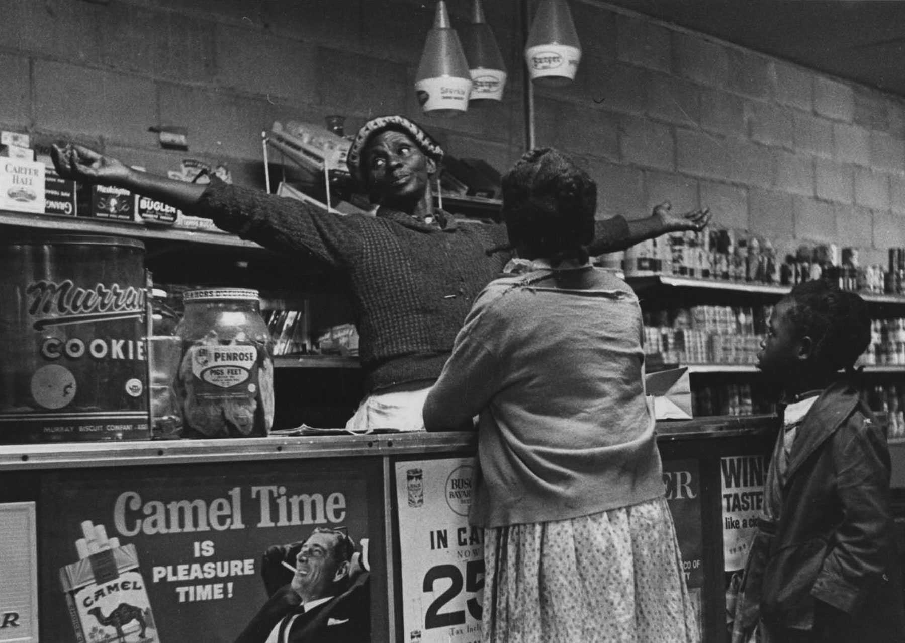 Behind the counter of the co-op, a woman stretches her arms out. Two young customers look at her from in front of the counter.