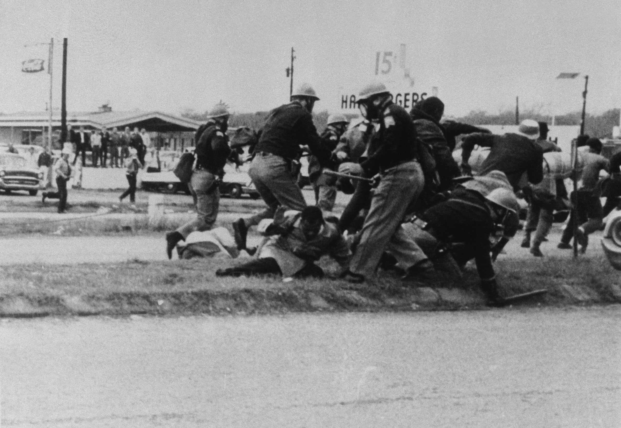 A black and white photograph of John Lewis trying to avoid a blow from a state trooper swings his club at Lewis' head.