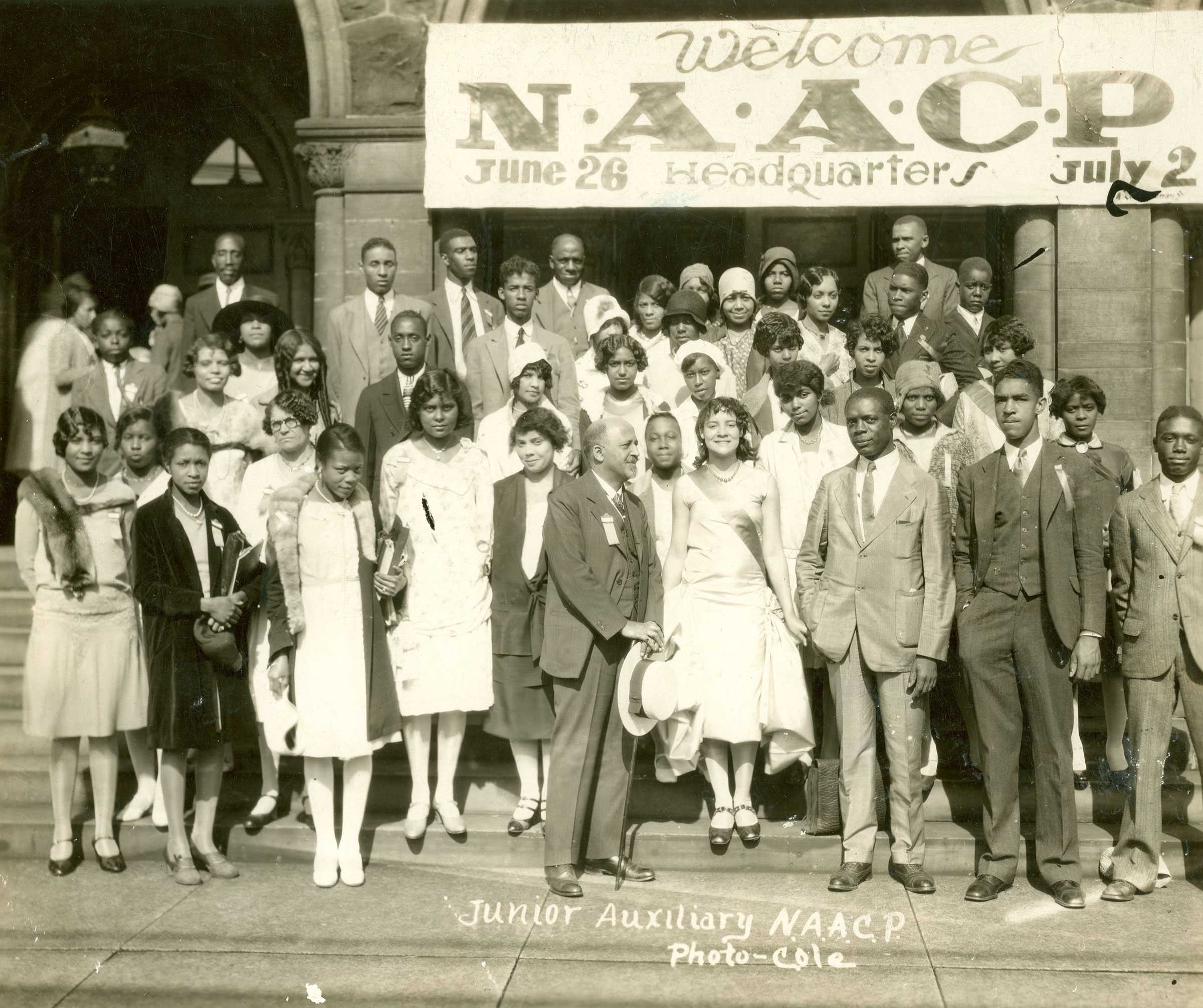 A group of people dressed in formal attire, stand on the steps of a building beneath a banner that reads 'Welcome N.A.A.C.P. June 26 - July 2 Headquarters.'