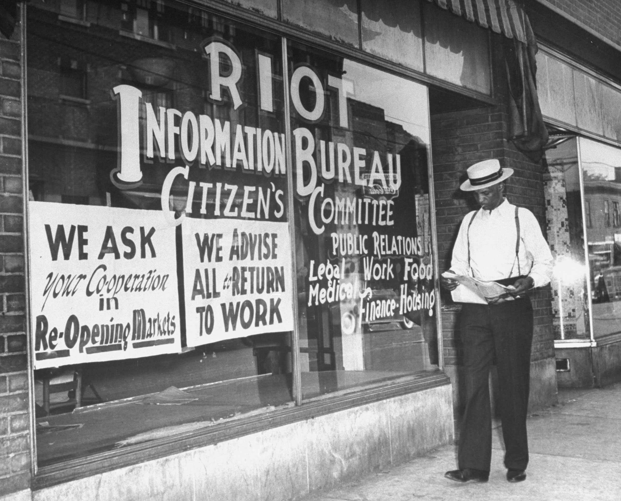 Black and white photograph of man walking and reading newspaper in front of store front marked" RIOT / Information Bureau / Citizen's Committee / Public Relations / Legal / Work / Food / Medical / Finance Housing"  Two additional signs hang in the window "WE ASK / your Cooperation in / Re-Opening Markets"  and "WE ADVISE / ALL to RETURN / TO WORK."