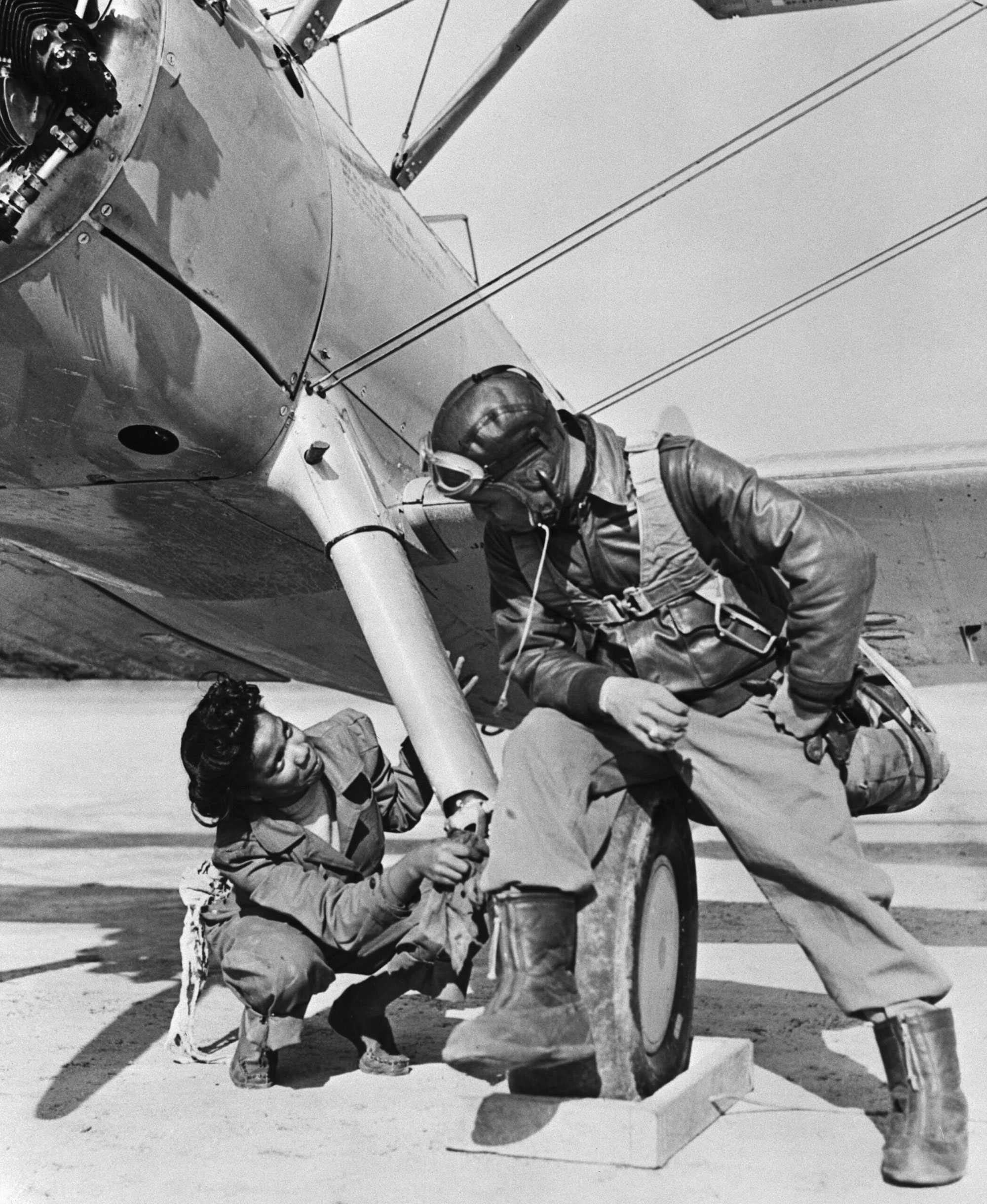 Black and white photograph of a African American woman mechanic working on a military plane.  She is squatted and looking up as a soldier speaks to her.