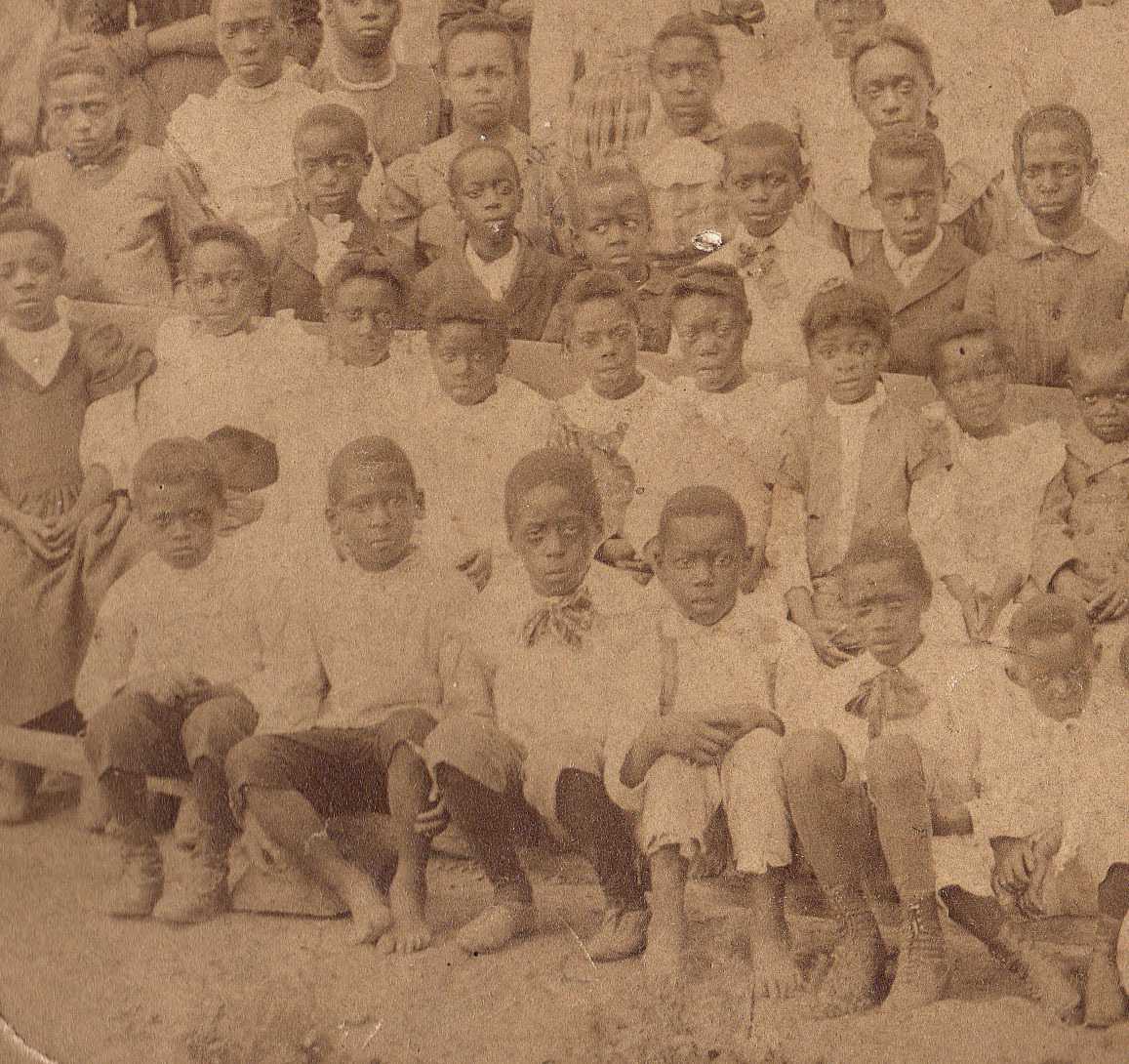 Photograph of African American children dressed in school uniforms and seated outside in three rows of benches.