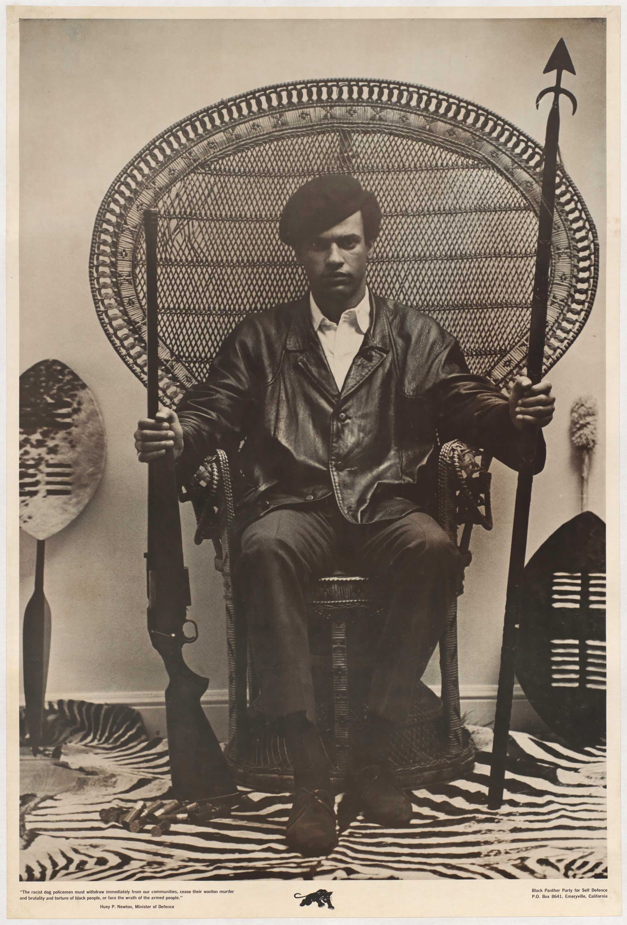 Man (Huey Newton) sitting in a rattan throne chair wearing a beret and a black leather jacket while holding a shotgun in his right hand and a spear in his left hand. Leaning against the wall on either side of the chair is a leaf-shaped, Zulu style shield with designs of horizontal line markings across the front.