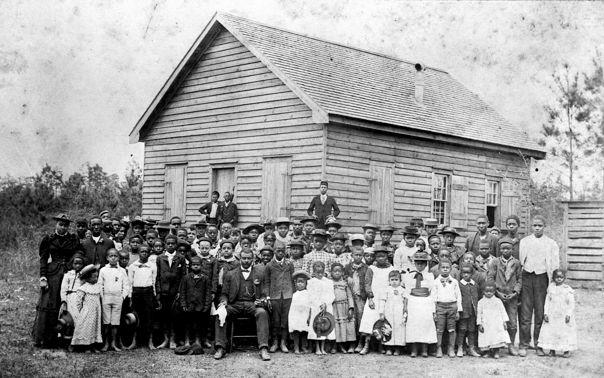 A group of African-American students and teacher stand in front of small log school.