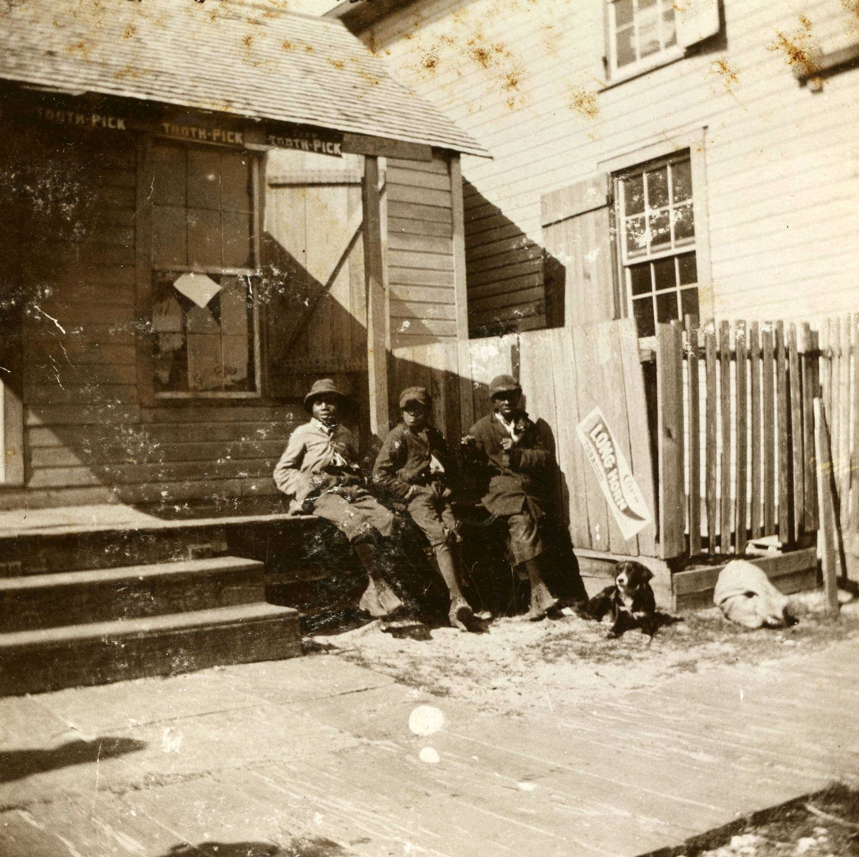 A worn sepia toned photograph of 3 boys sitting on the steps of a store. Each have their hands in their pockets.