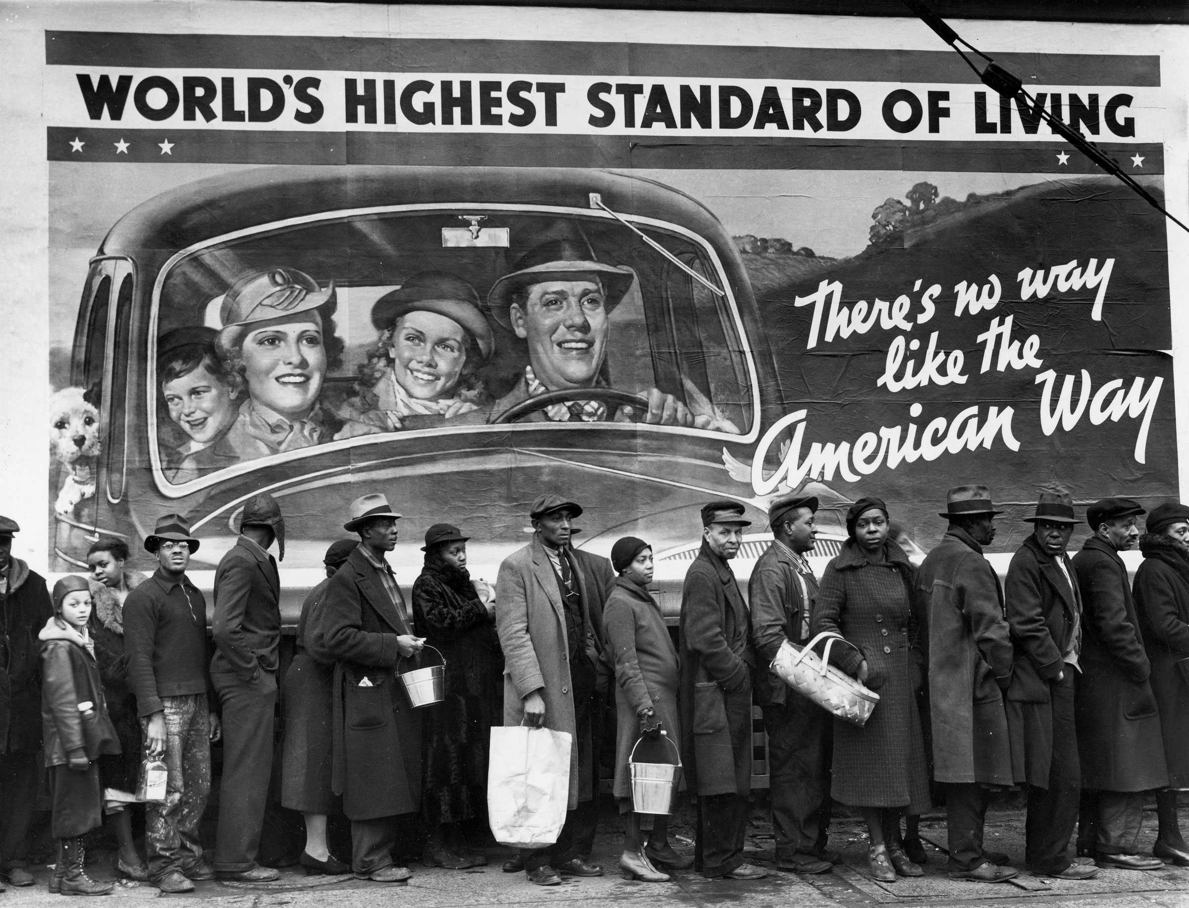 Black and white photograph of a line of African American people lined up in front of a billboard .  They are dressed in winter clothing.  The Billboard entitled " WORLD'S HIGHEST STANDARD OF LIVING"  depicts a carefree caucasian family driving in a car.  The billboard also says "There's no way like the American Way."