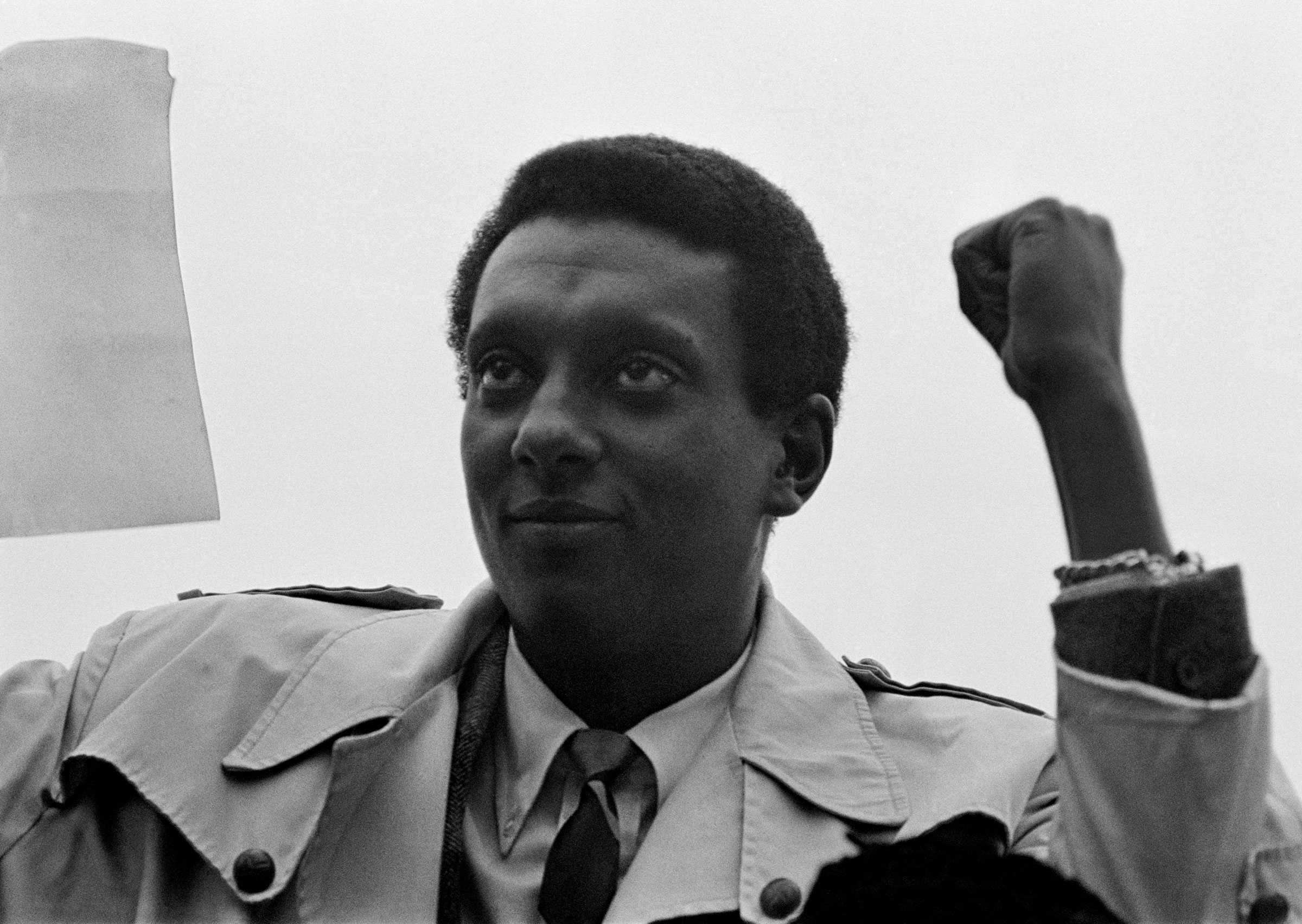 Stokely Carmichael holds a paper and his fist in the air at a peace rally. Dressed in a suit and raincoat, he looks forward towards the crowd.