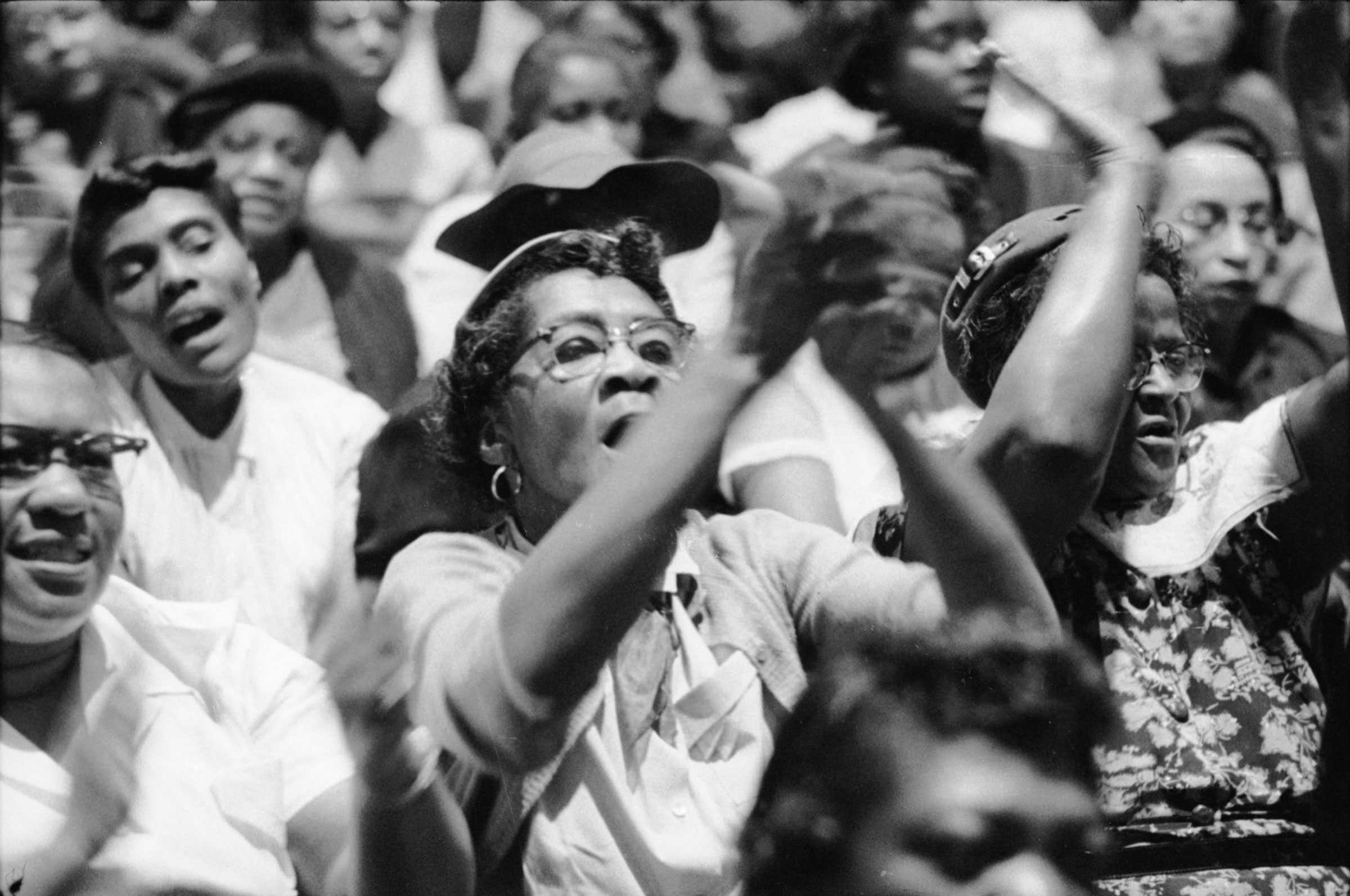 A black and white photograph of a crowd cheering and clapping at a Montgomery Bus Boycott Meeting.