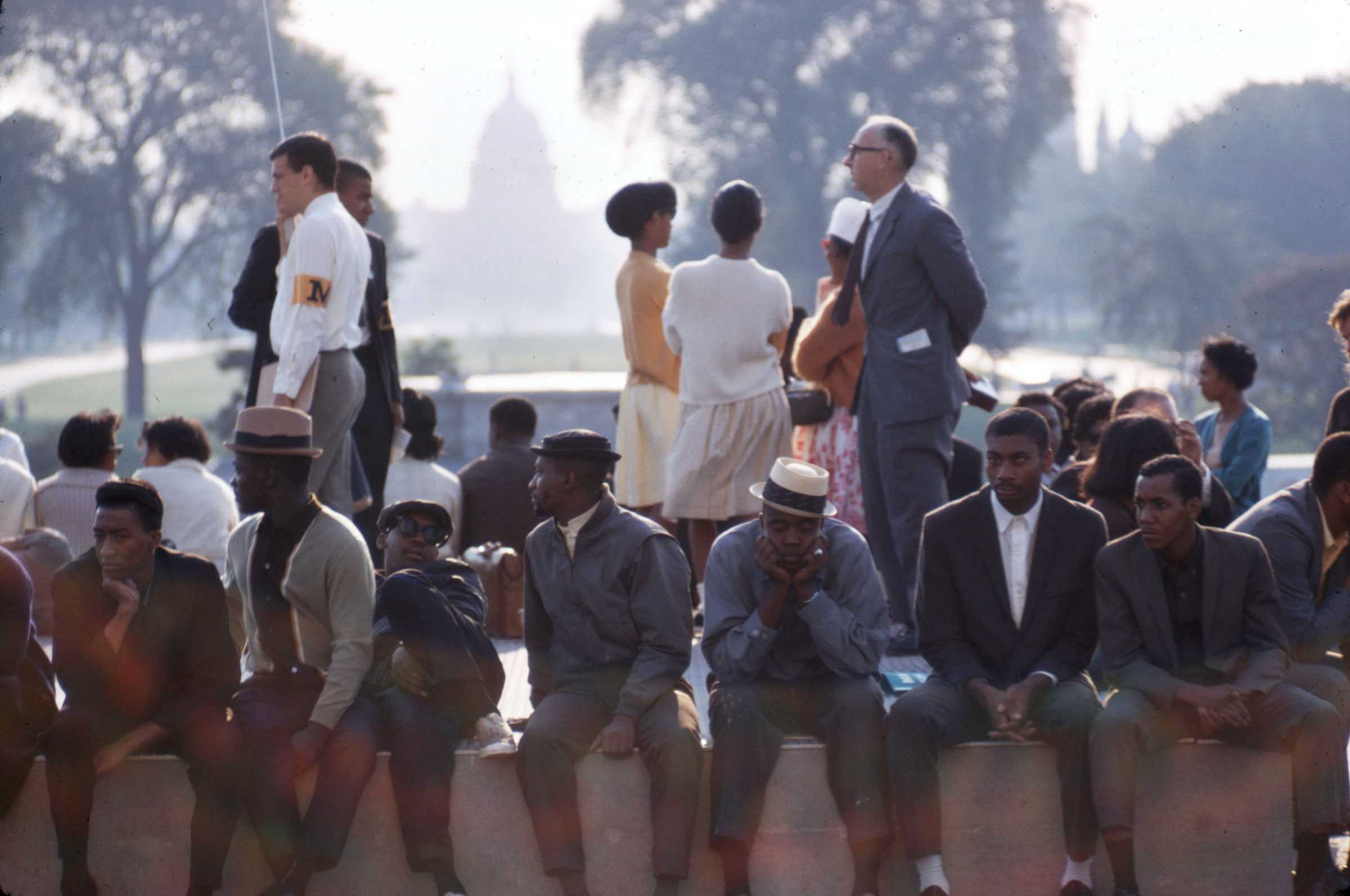 March on Washington participants seated on a ledge with others standing behind them, and U.S. Capitol in the background. Trees frame the scene.