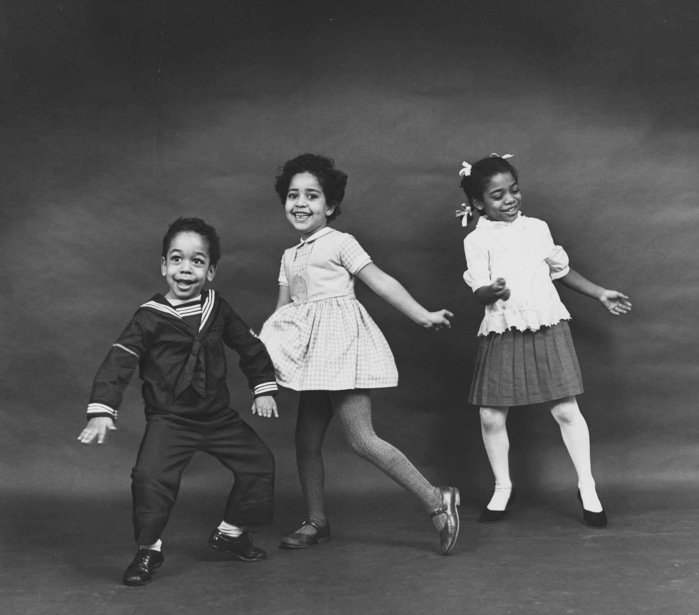 A black and white photo of three children dancing: a boy in a sailor suit, and two girls in dresses.