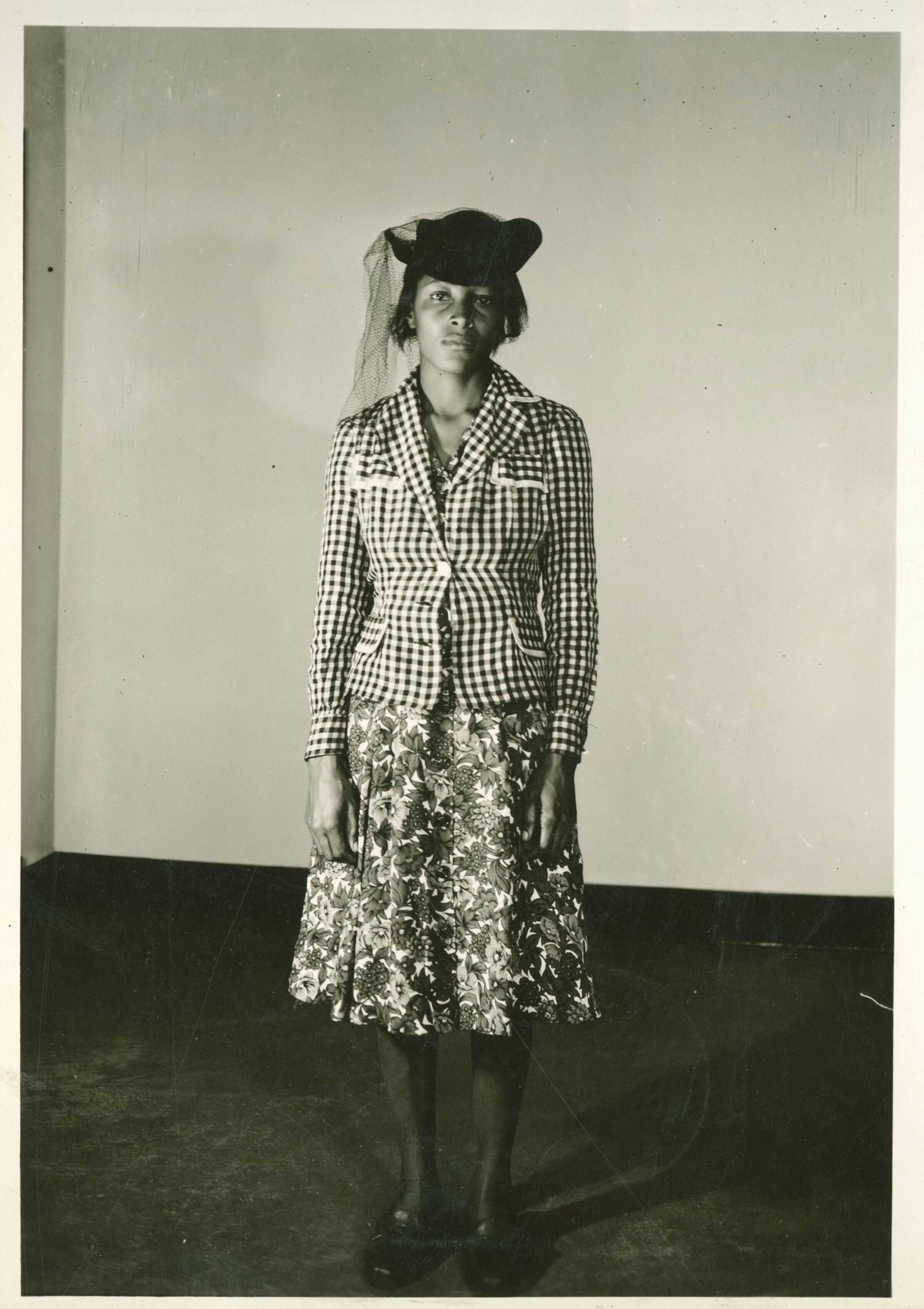 Recy Tailor stands in a for a full body sepia toned portrait. She is wearing a plaid blazer, a flower skirt, and a black hat.