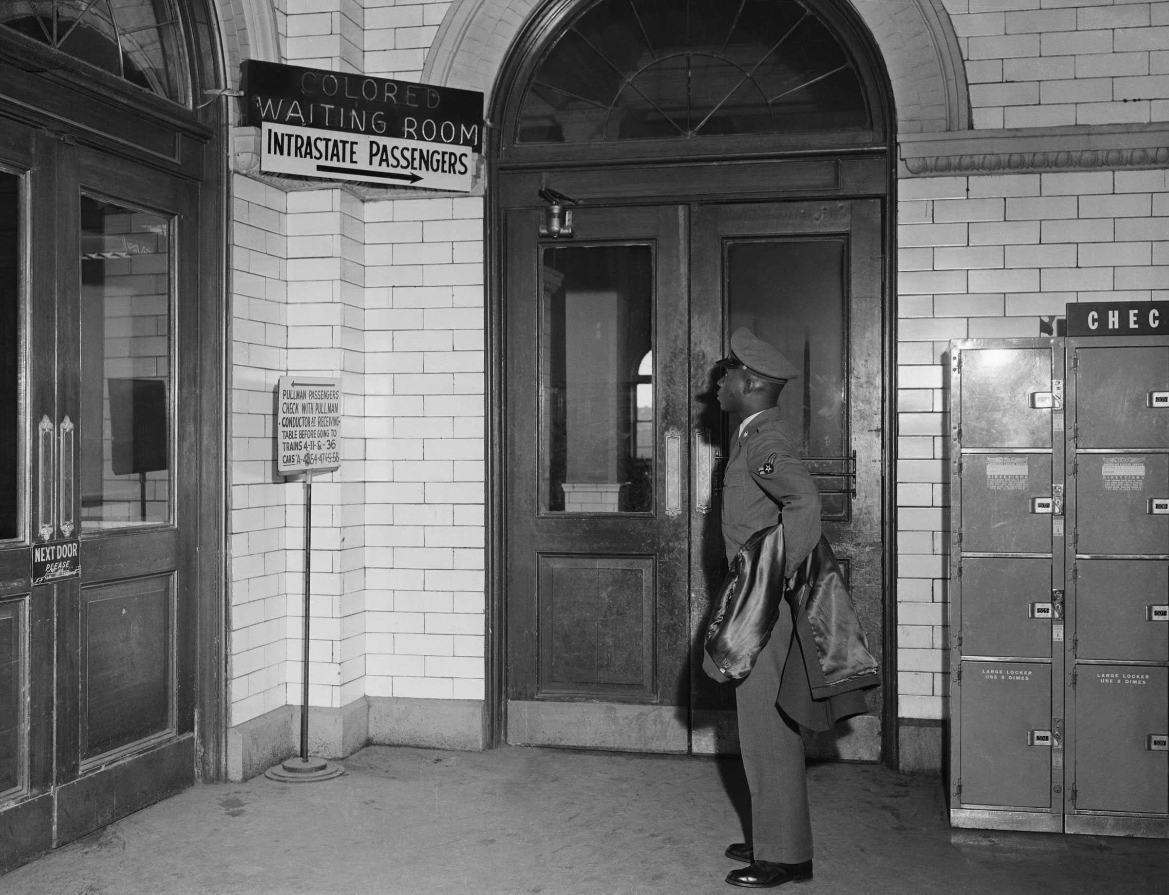 Black and white photograph of Black man dressed in military uniform reading a sign in a terminal marked "COLORED/ WAITING ROOM/ INTRASTATE PASSENGERS."