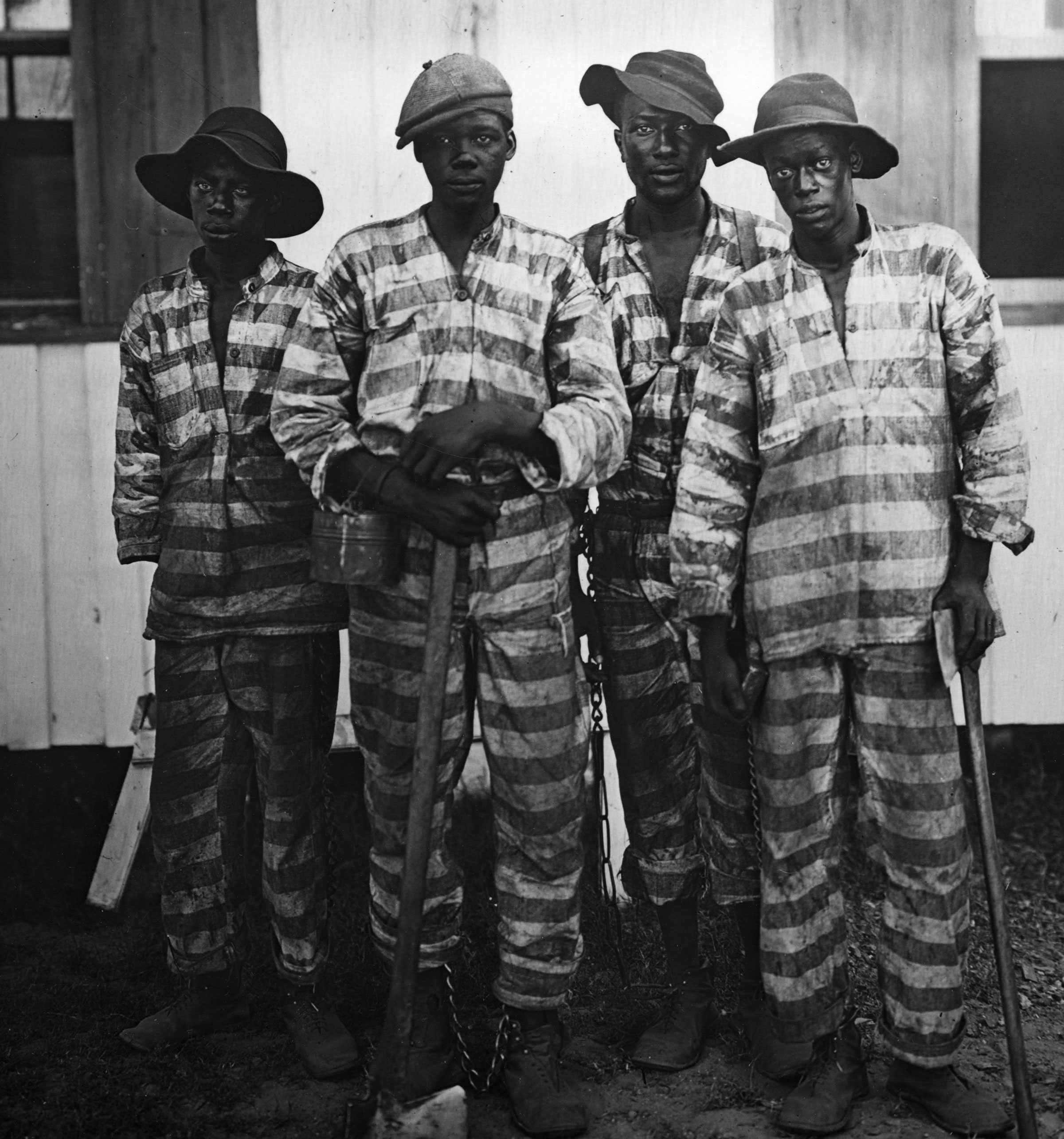 Black and white photograph of 4 african american men dressed in striped prison clothes holding axes and other tools.  There are chains visible around their ankles.