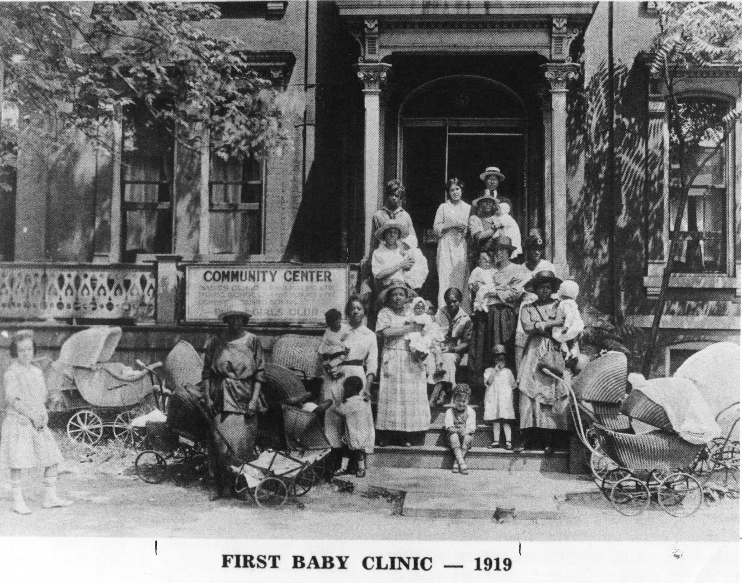 Black and white photograph of women and children standing on the front steps of a house.  There are baby carriages on both sides of the women.  The Photograph is titles "FIRST BABY CLINIC - 1919.