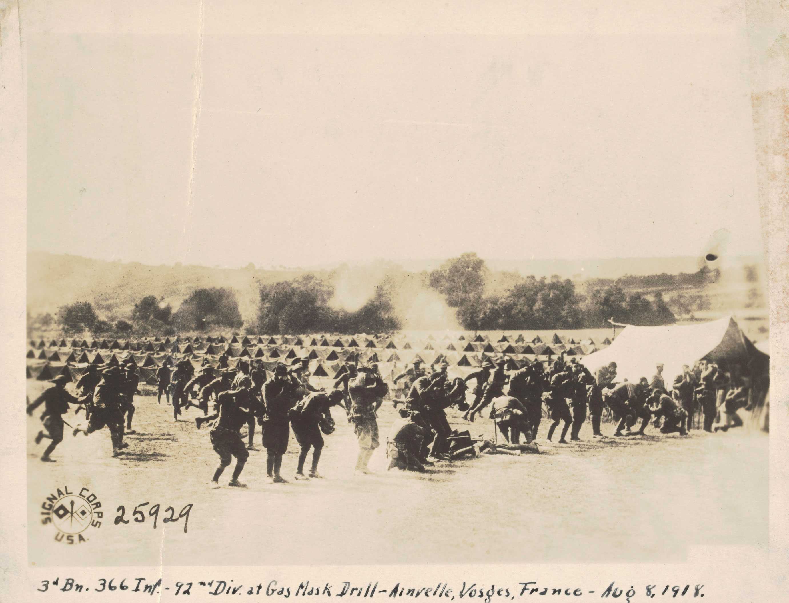 A black and white photograph of soldiers participating in a gas mask drill. The soldiers are standing, running, and kneeling as puffs of gas fill the air.