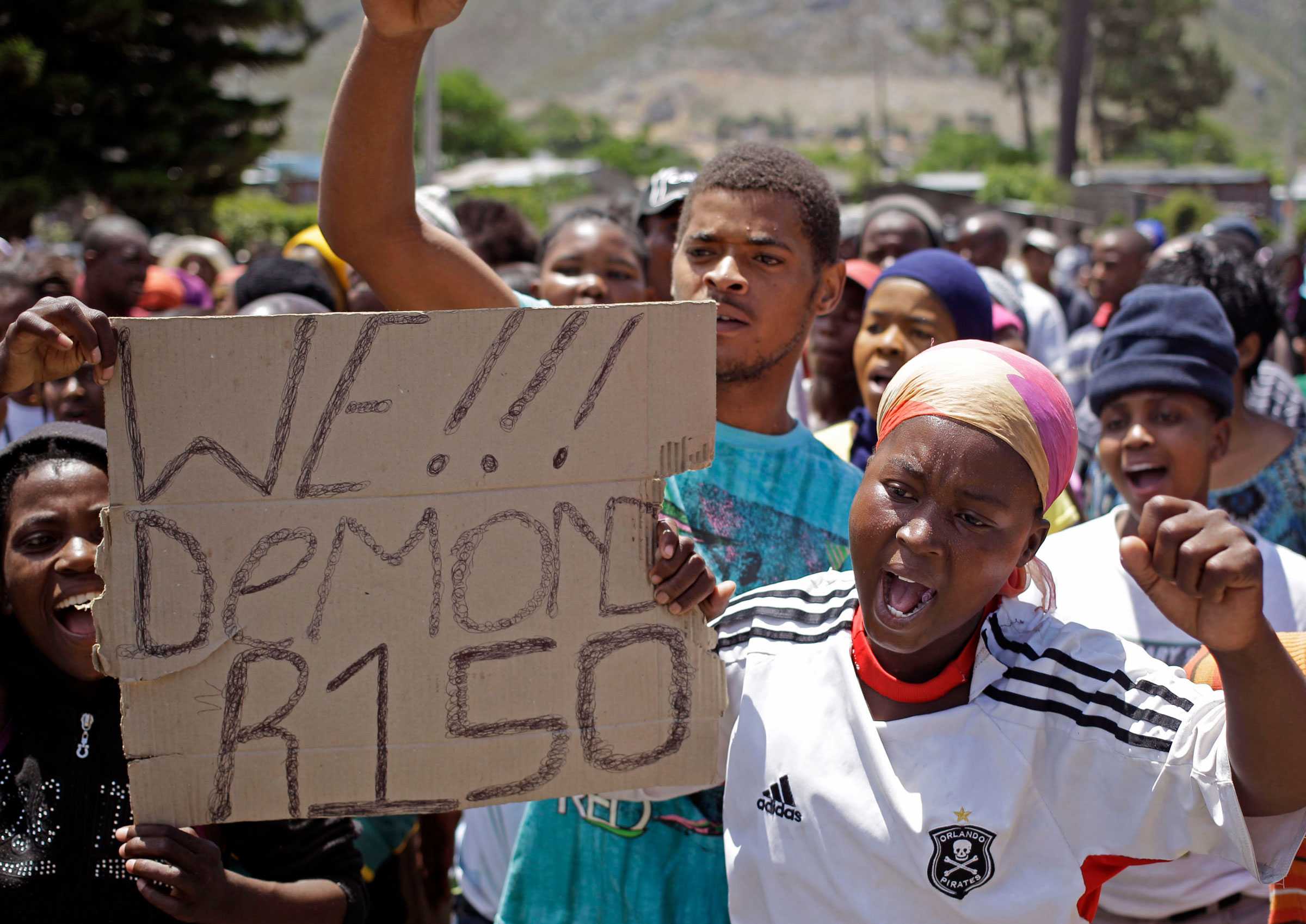 Farm workers chant and protest. One woman is holding a cardboard sign that says 'We!!! Demond R150'