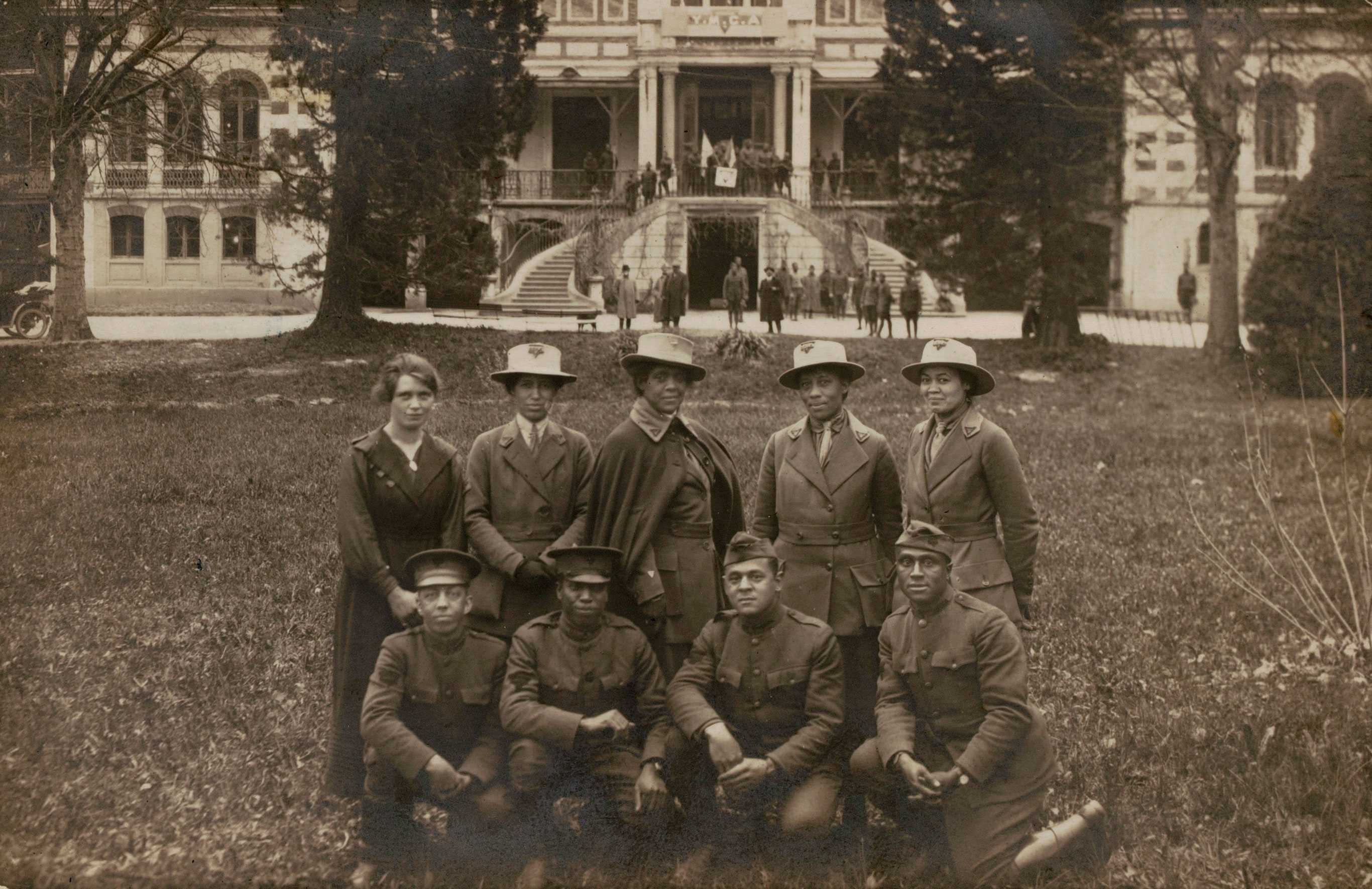 A real photo postcard of a group of military service men and women taken at the YMCA camp near Chambery, France, during World War I. The image depicts five women standing in a row on a lawn, with four men crouched in a row in front of them. Addie Waites Hunton is in the center of the back row; the other women and men are unidentified. In the background is a large building with a double staircased entrance. A temporary sign reading [Y.M.C.A.] has been placed on the portico at the top of the stairs. Other individuals are visible along the top and bottom of the stairs. The verso has printing reading [CARTE POSTALE] with spaces for [Correspondance] and [Adresse] and a horse and horsehead mark for the publisher Guilleminot. The postcard has not been sent, but there is an inscription across the back by hand in brown ink reading [From Sgt. Thomas, who / was on leave at colored, / Y.M.C.A. at Chamberry / France]. There is an inscription by a different hand in graphite above the [Adresse] label reading [(ALFRED JACK THOMAS)].