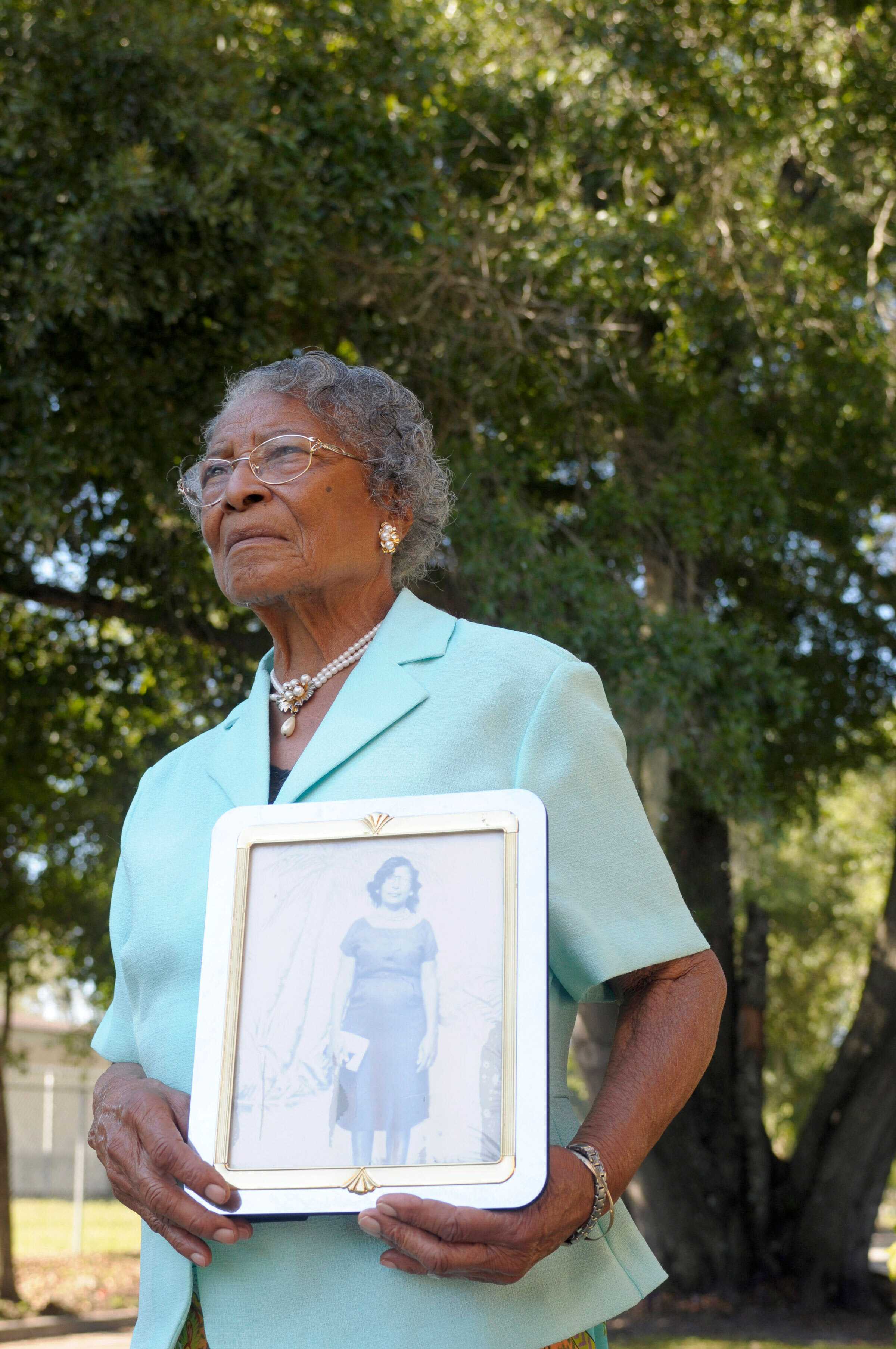 Color photograph of an African American woman dressed in a pale blue shirt holding a framed photograph.