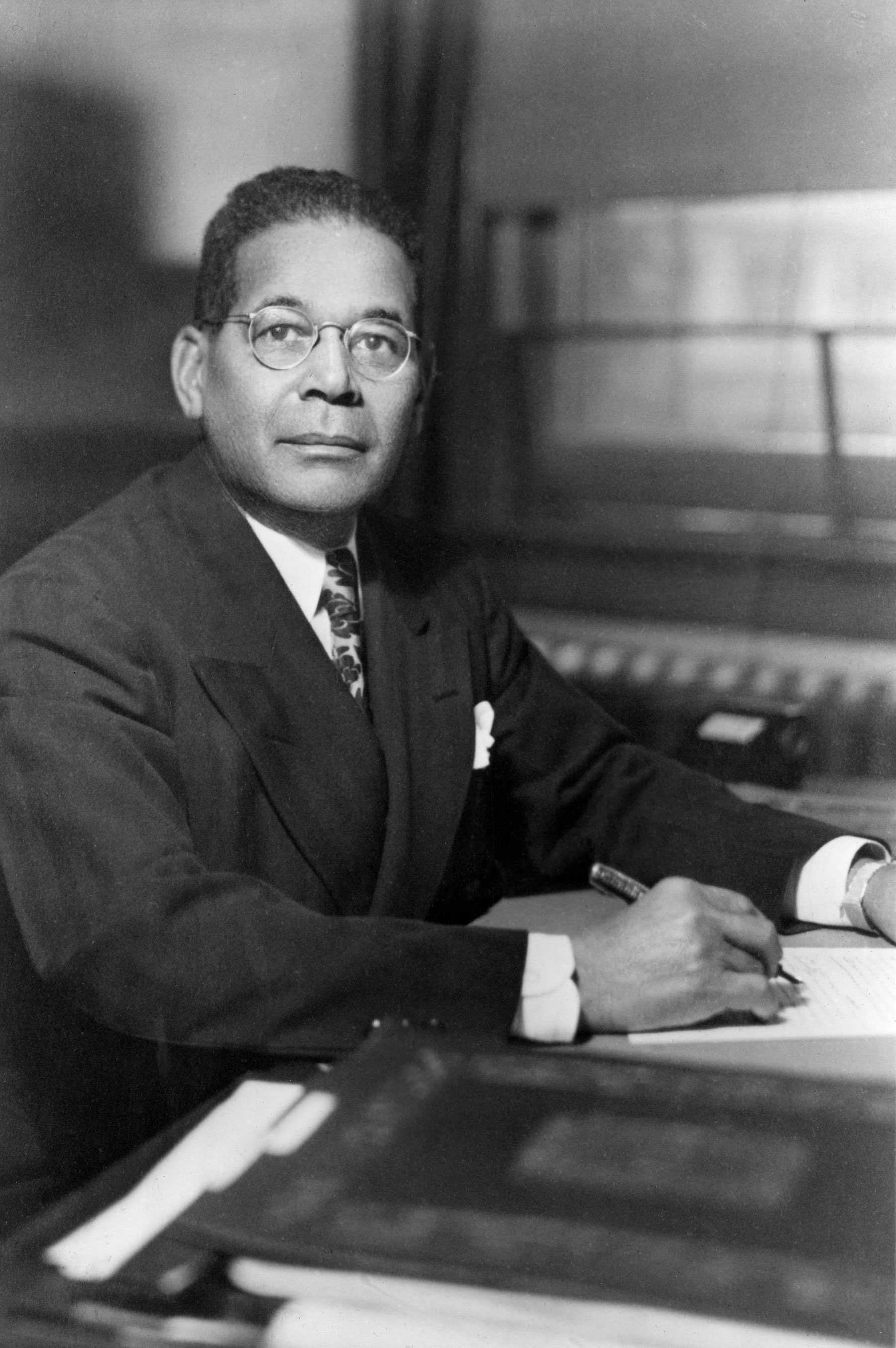 Photograph of a man seated at a desk writing on a pad of paper.  The man is dressed in a suit and tie with a white shirt.  He is also wearing glasses as he looks up at the viewer.