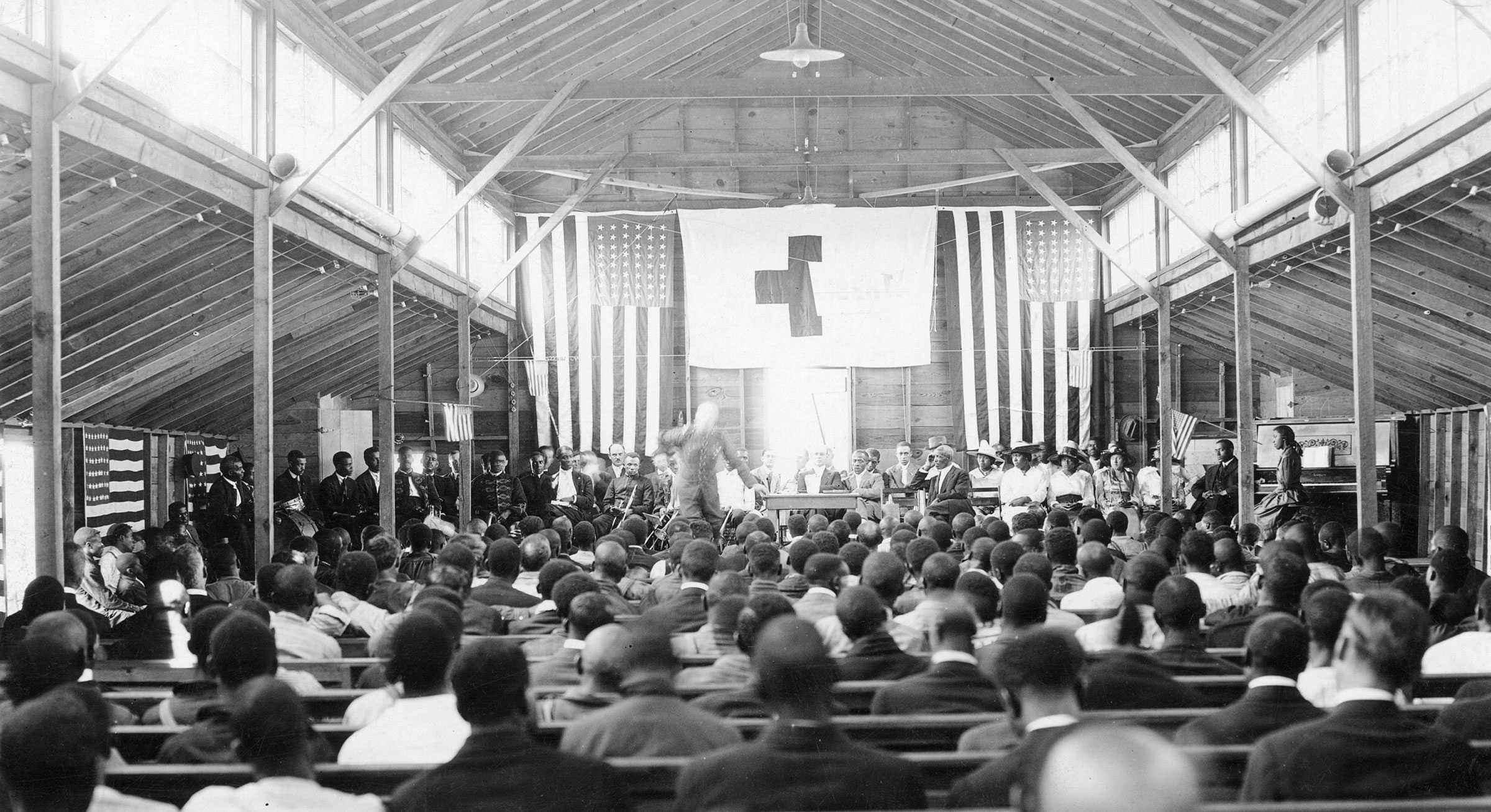 Black and white photograph taken from the back of a large hall.  The hall is full of people and there are three flags hung at the front of the hall including two American Flags and a third with a cross on it.