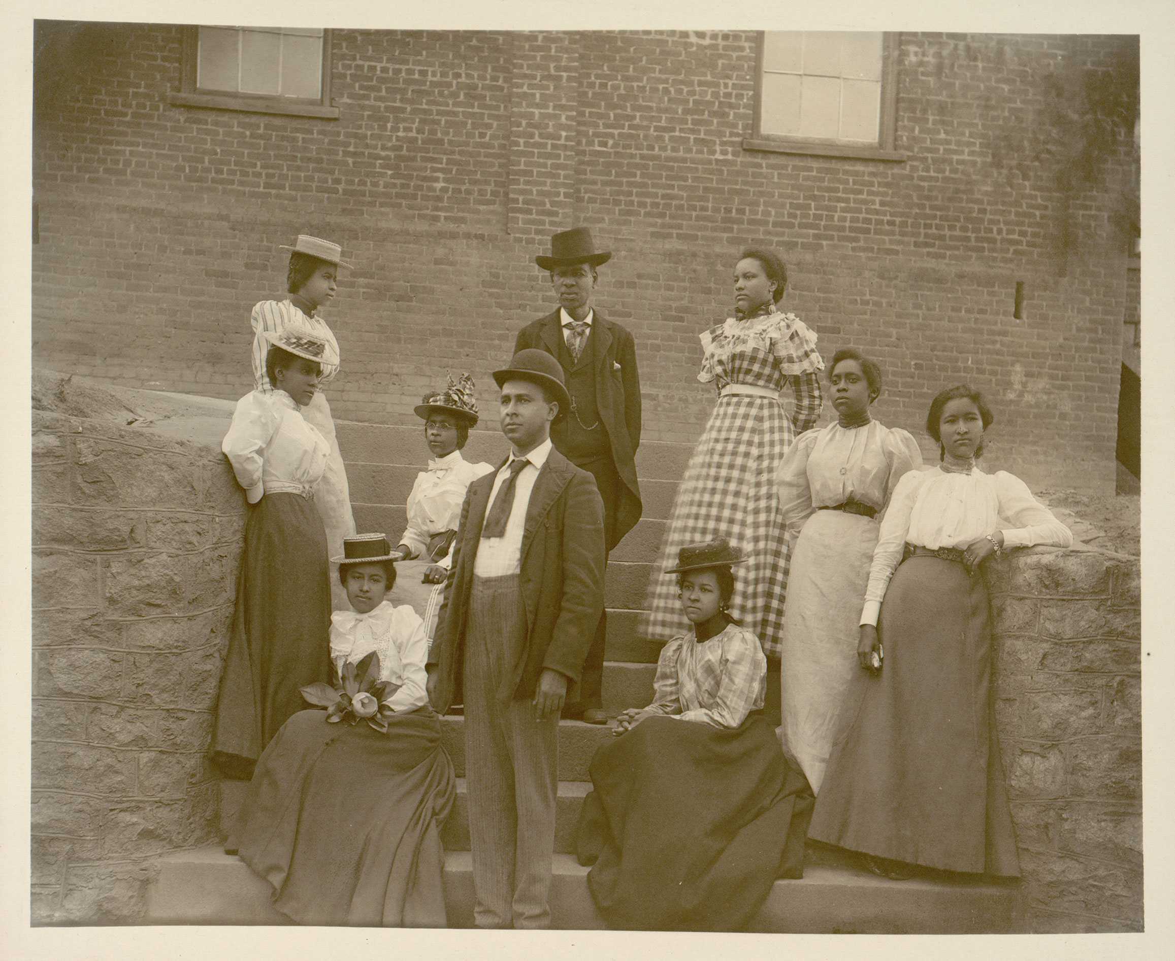 A sepia toned photo of a group of women and men posing for an image. Each are dressed up with long dresses, suits, and hats.