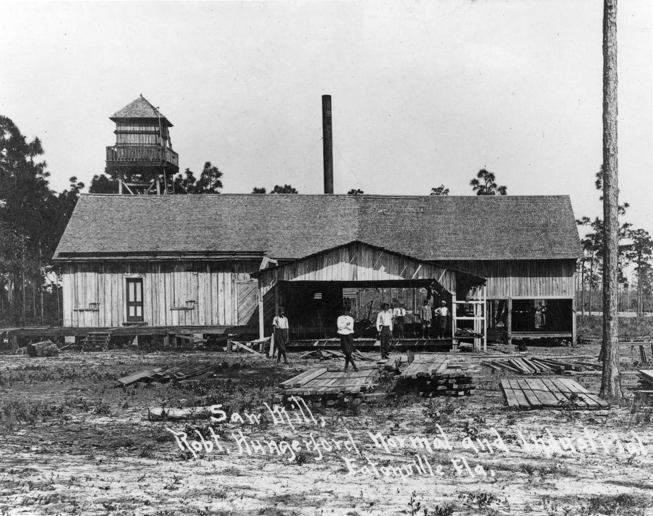 A black and white photograph of the sawmill. Three balck men stand in front of the building.