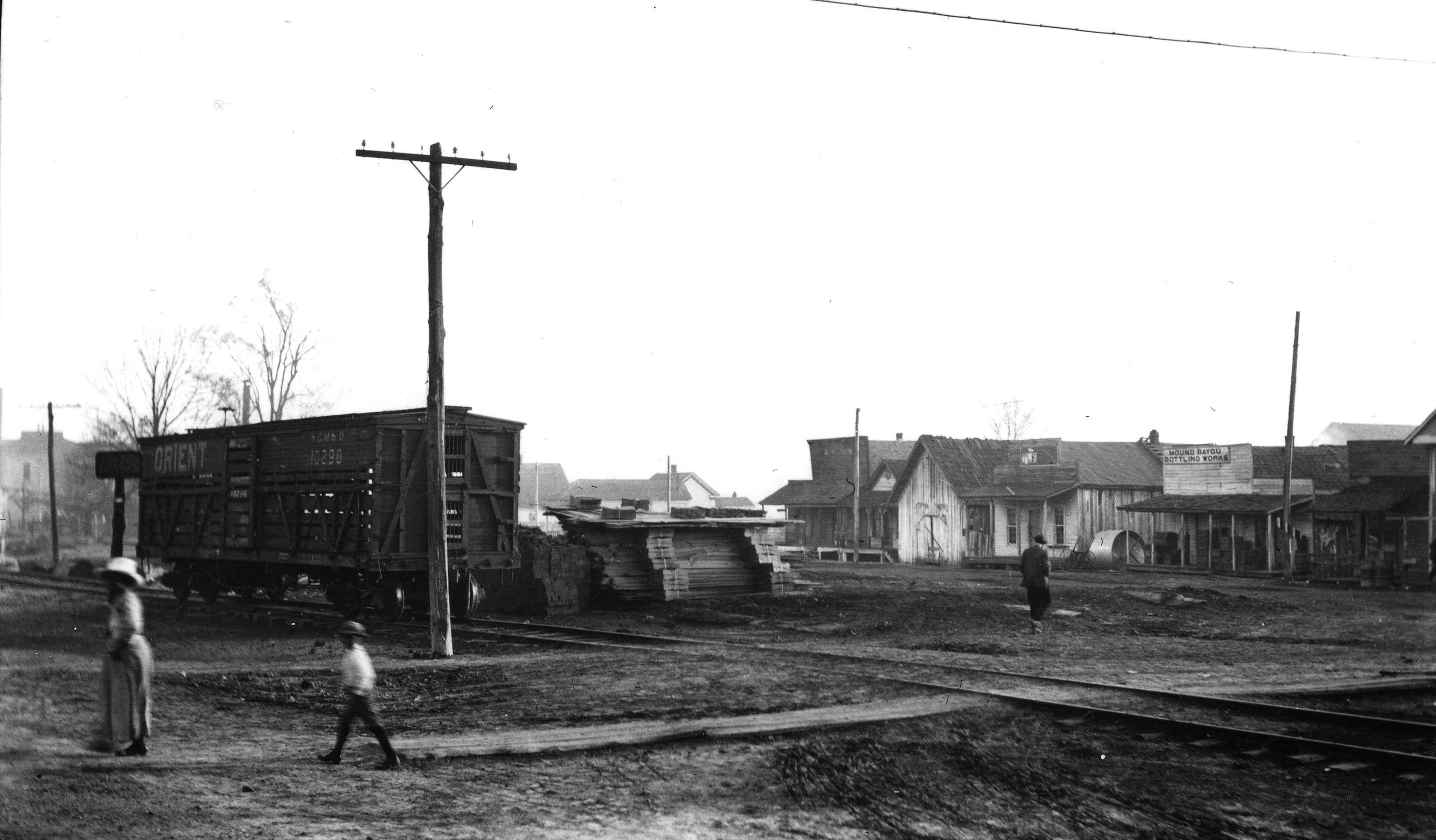 A landscape view ot Mound Bayou's buildings and town. Three blury figures walking across the dirt paths.