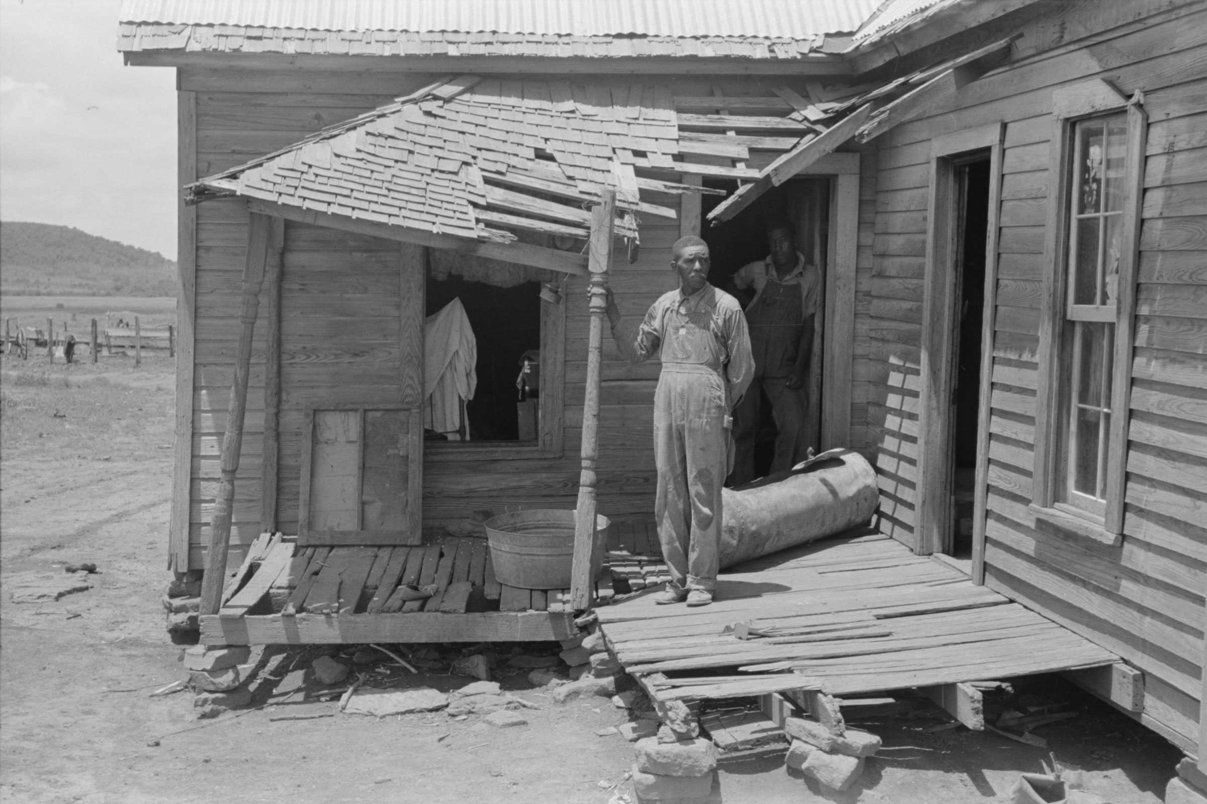 Black and white photograph of an African American man standing on the porch of a dilapidated wooden house.  The porch and roof are caving in.  There is another man in the background standing in the doorway.  Both men are dressed in overalls and shirts.