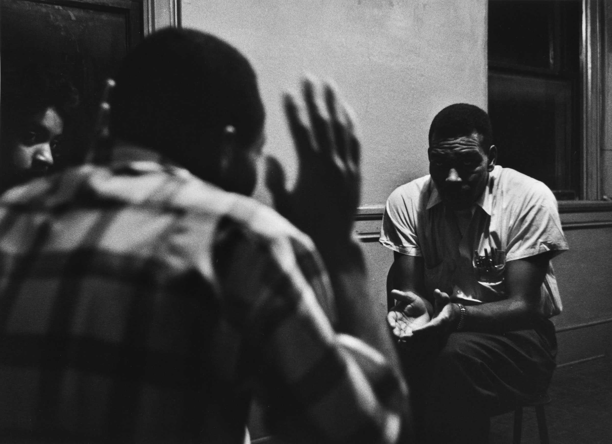 Black-and-white image of two men and a woman in discussion. One man, wearing a plaid shirt, has his hands raised, while the other man leans forward, hands clasped.