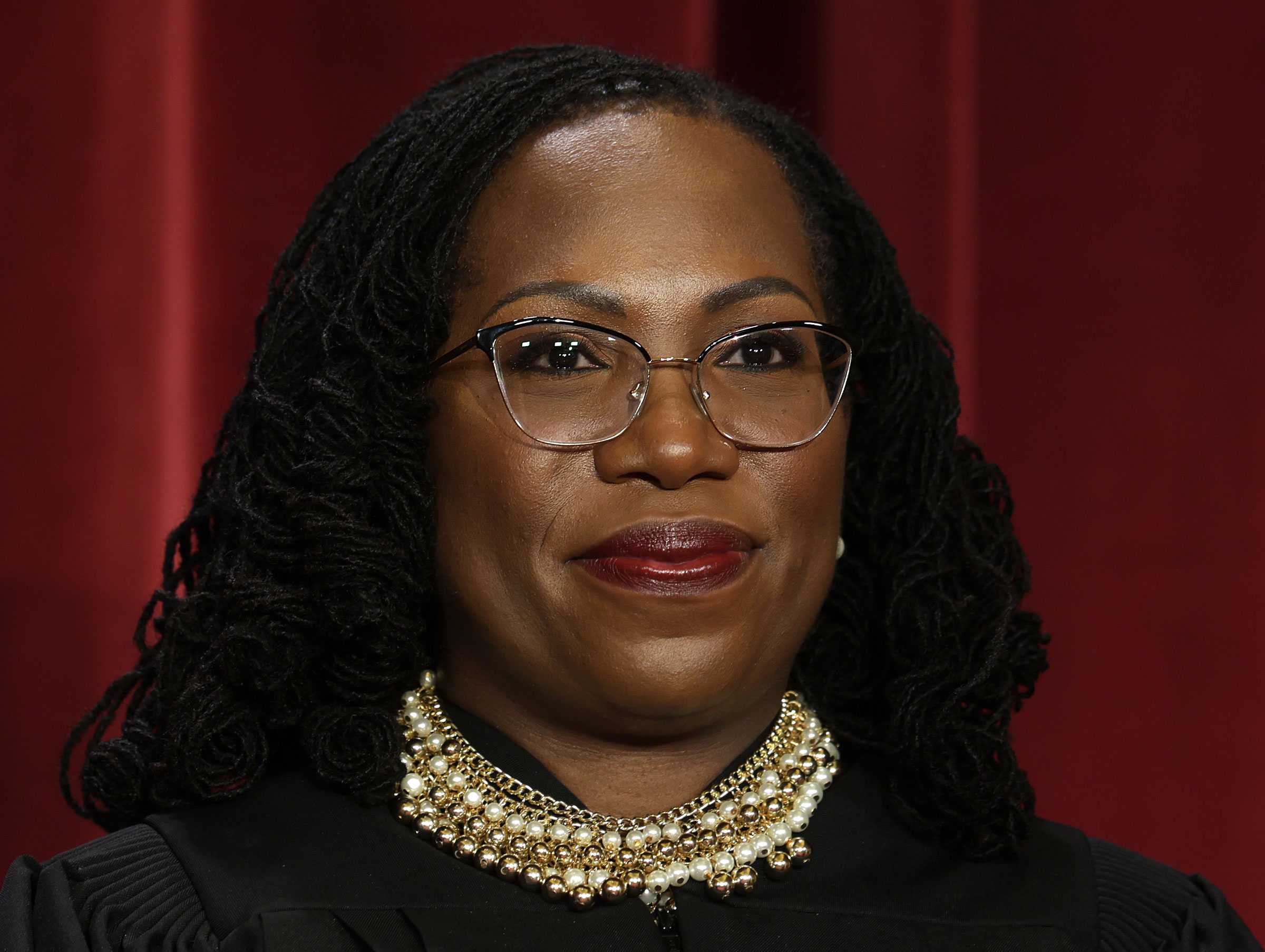 A close view of Justice Ketanji Brown Jackson. In front of red curtains, she looks off camera. She is wearing a large pearl and gold necklace.