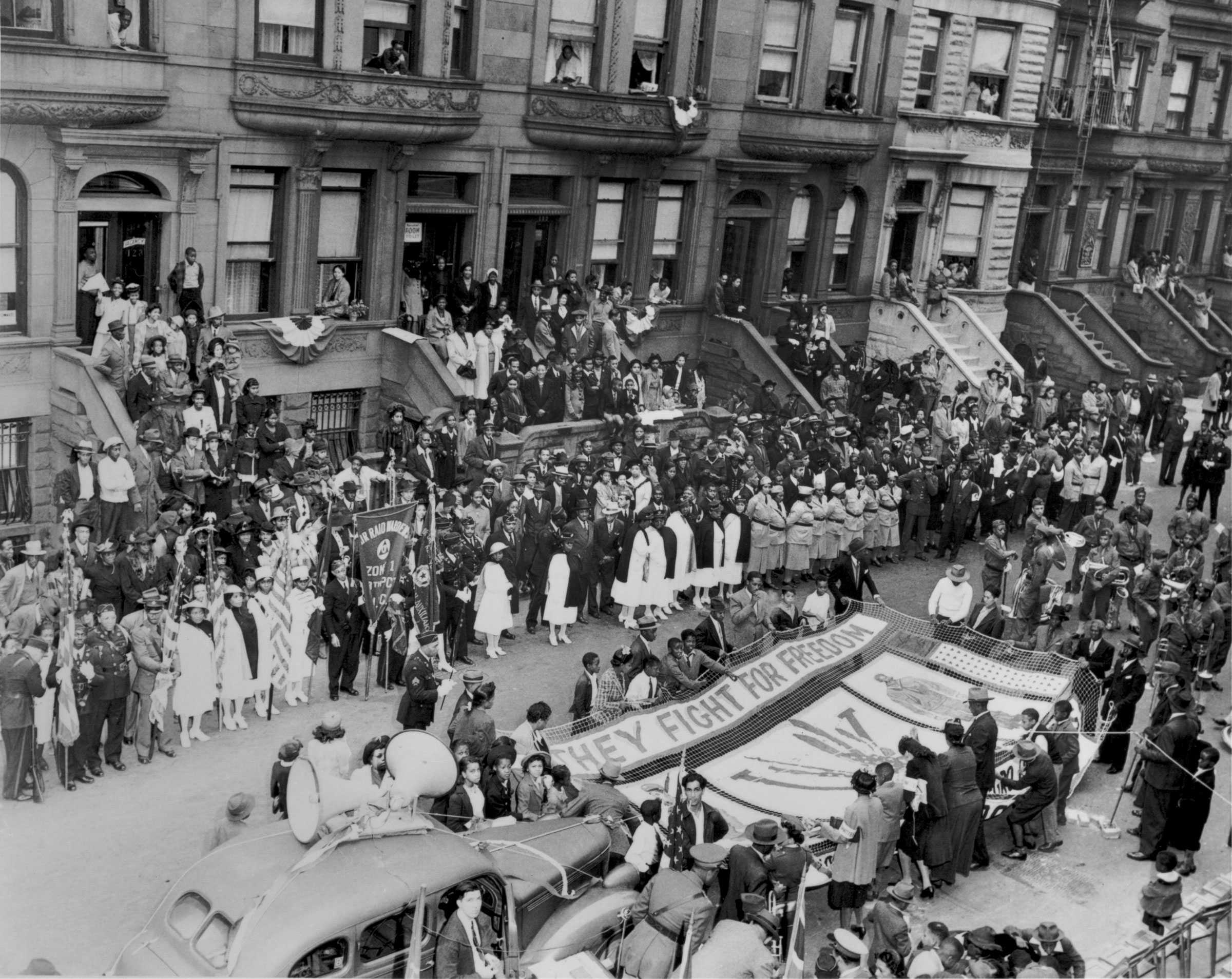 Black and white photograph showing a patriotic rally in an urban setting.  People are in the street and standing on the steps of buildings.  One group holds a banner reading "THEY FIGHT FOR FREEDOM / VV."