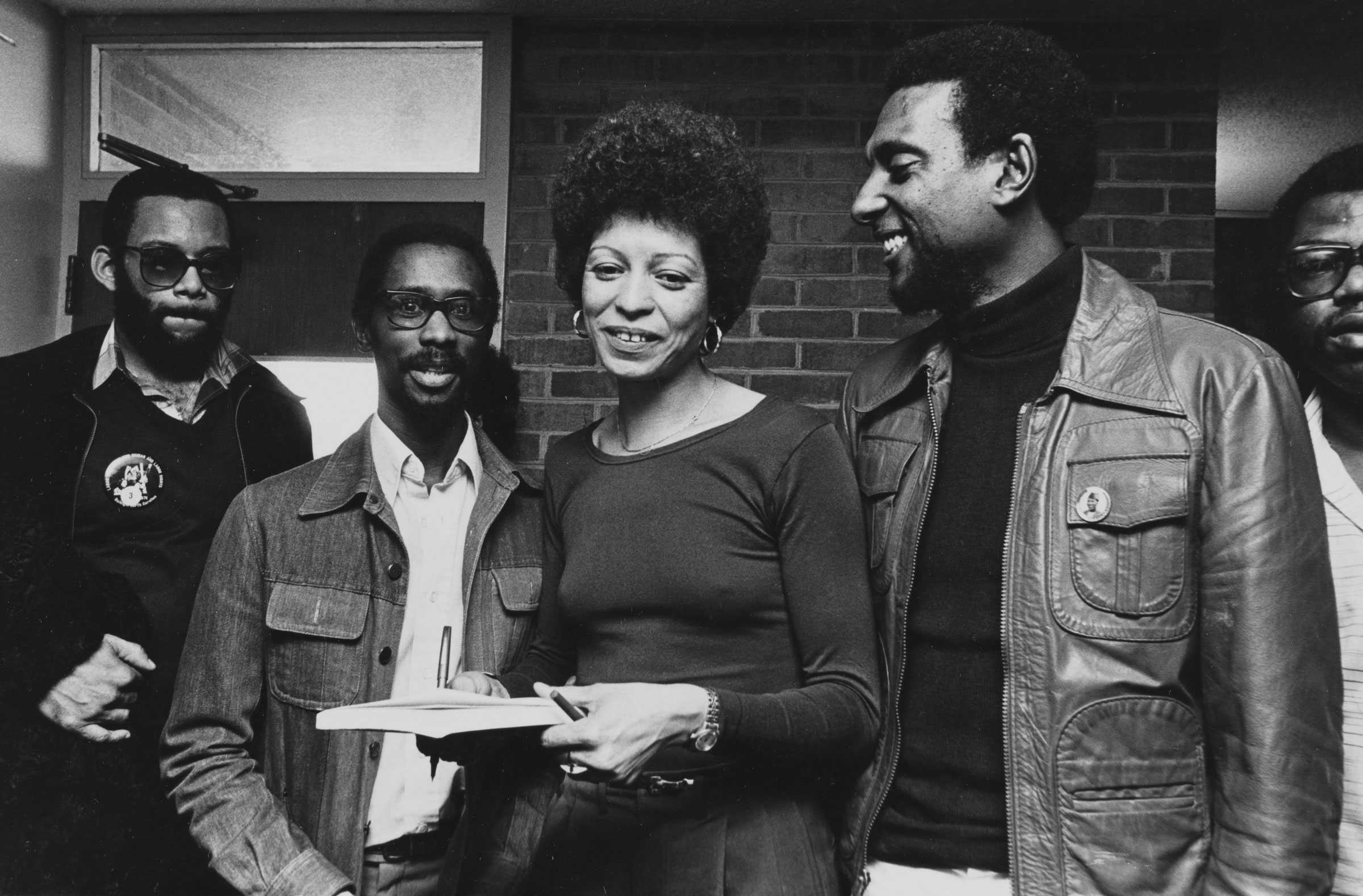 A black-and-white photograph of Angela Davis standing Stokely Carmichael, Jimmy Garrett, and Reginald Petty. Davis is holding a pen and book.