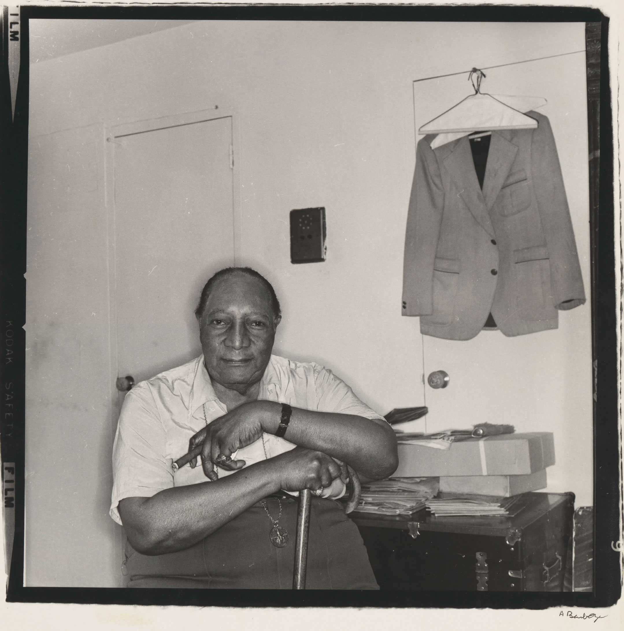 James Van Der Zee seated, holding a cane and cigar. A jacket is hanging on a door in the background.