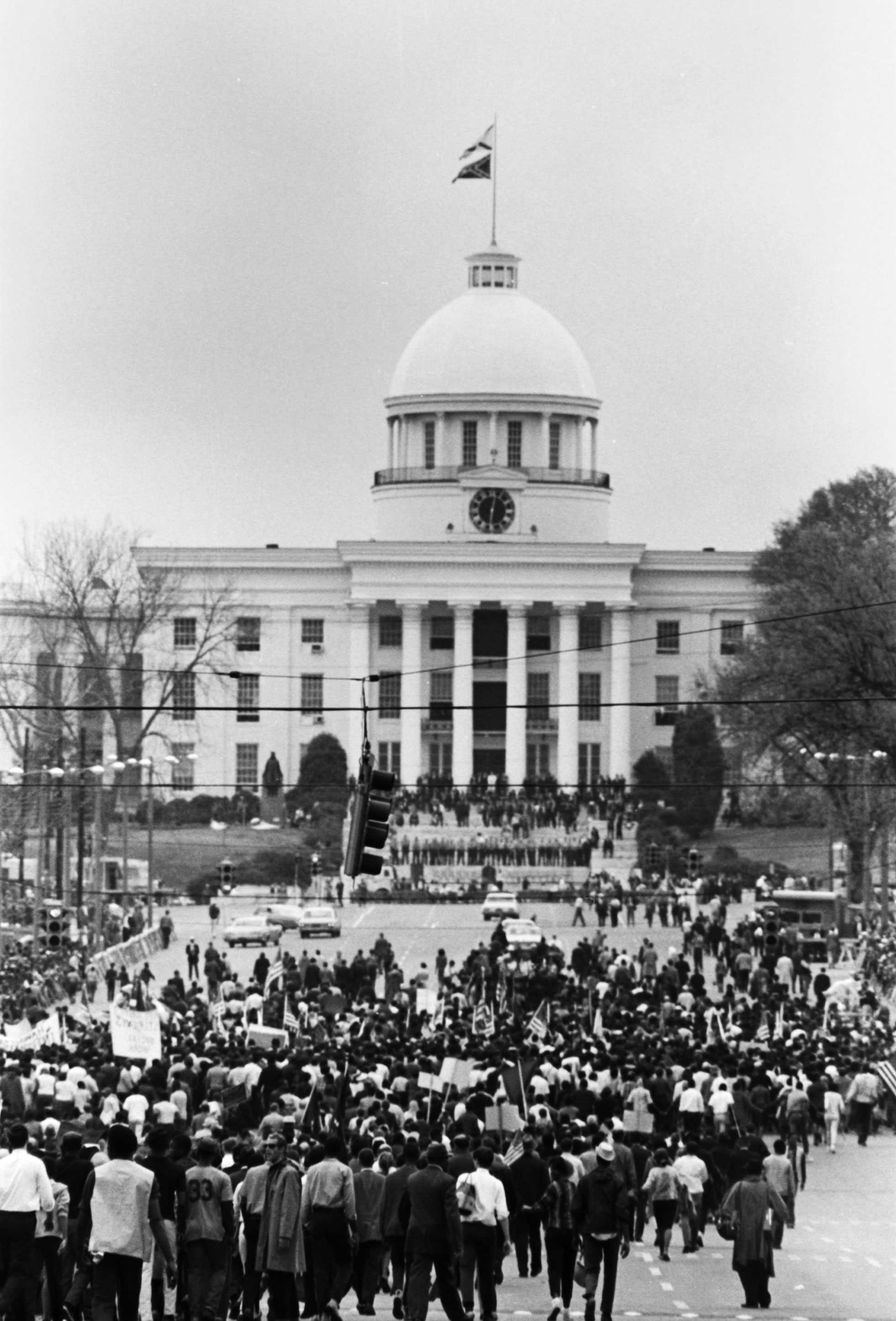 A black-and-white digital image of marchers walking toward the Alabama State Capitol. Marchers are seen from behind and are carrying signs and American flags.