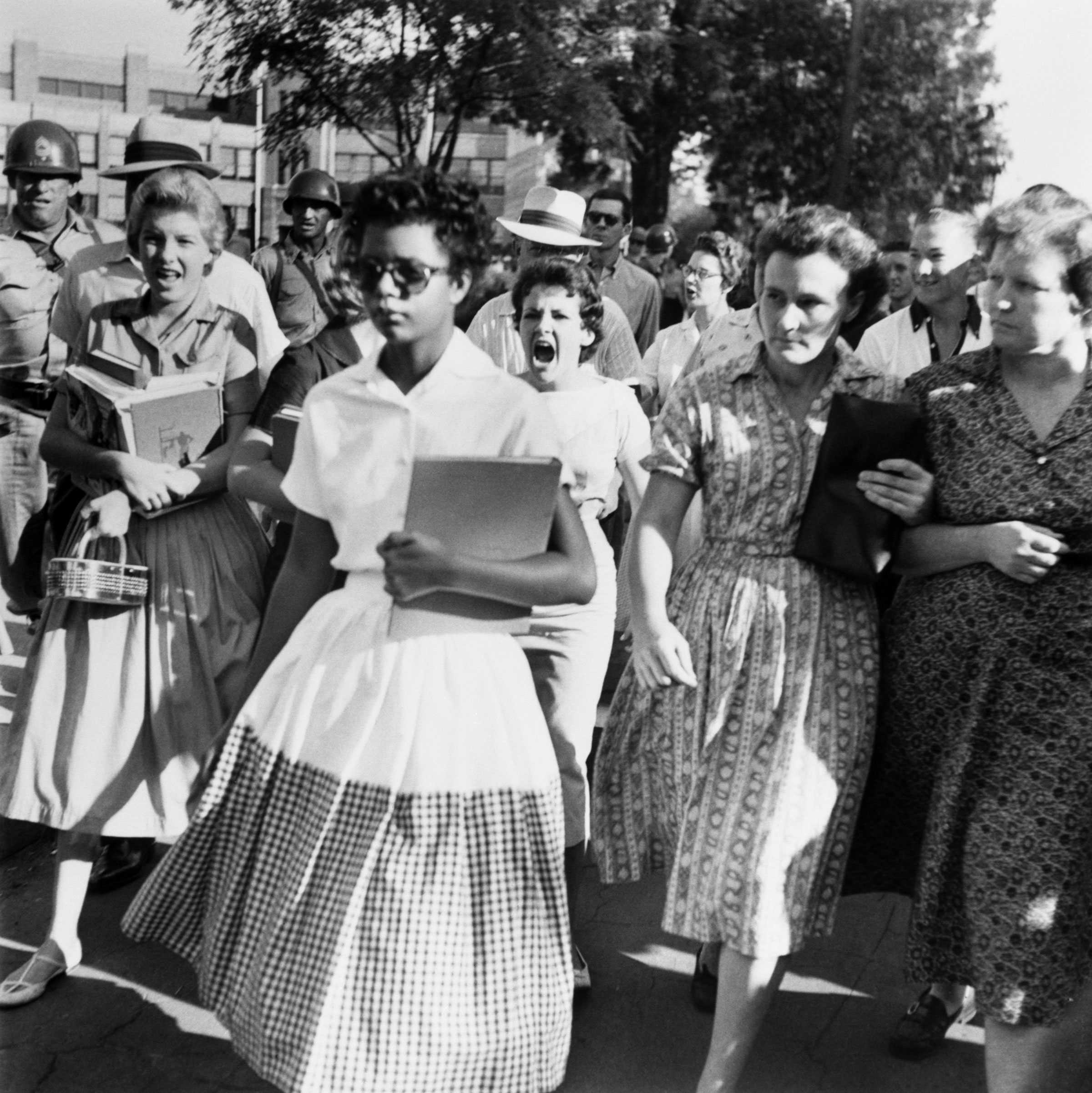 Elizabeth Eckford walks in a white dress and sunglasses as she ignores the hostile screams of women and students following her.