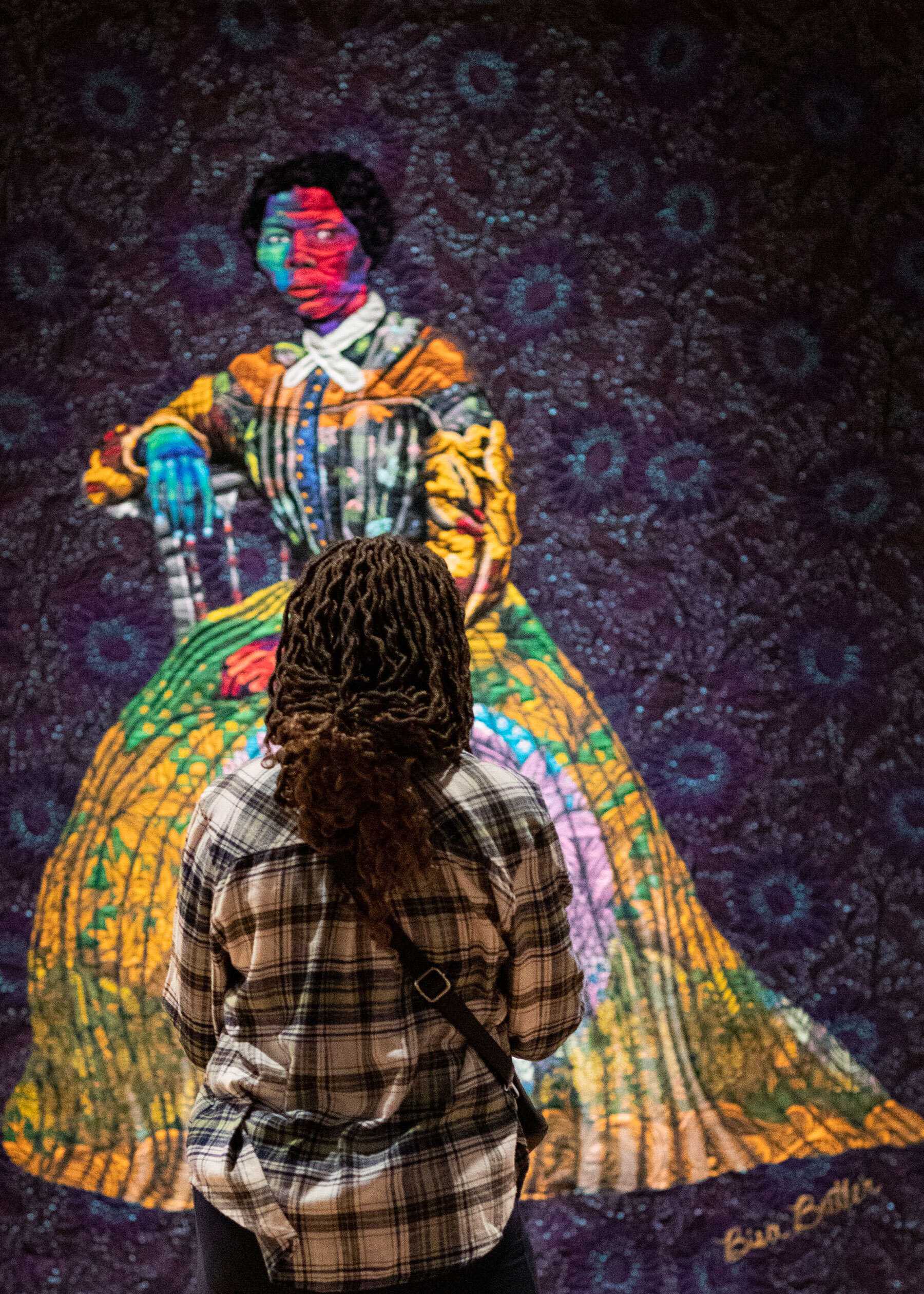 A view from the back by a woman who is looking at a quilted textile portrait of Harriet Tubman, made by Bisa Butler.