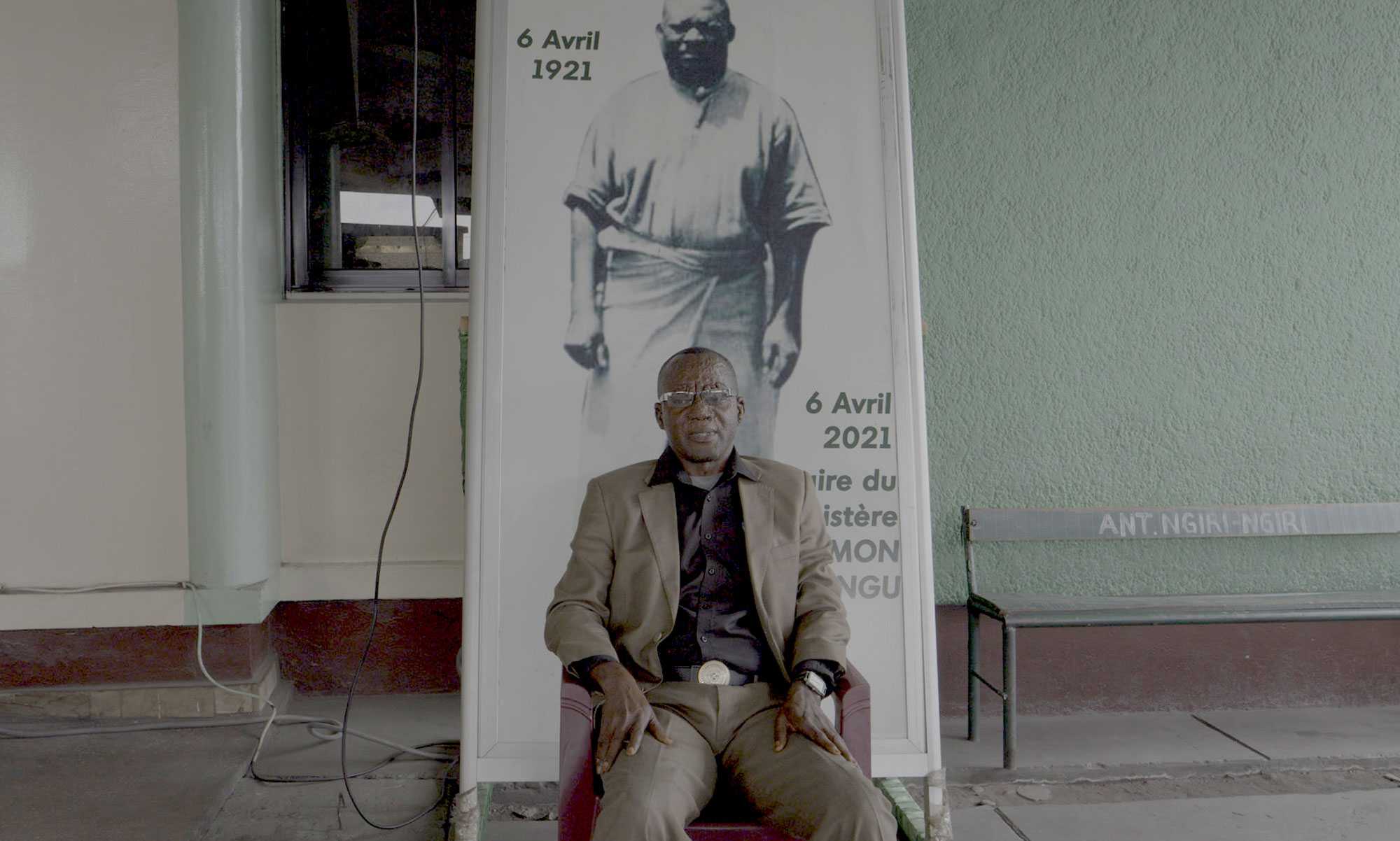 An older man in a light brown suit sits in front of a large photo of his younger self.