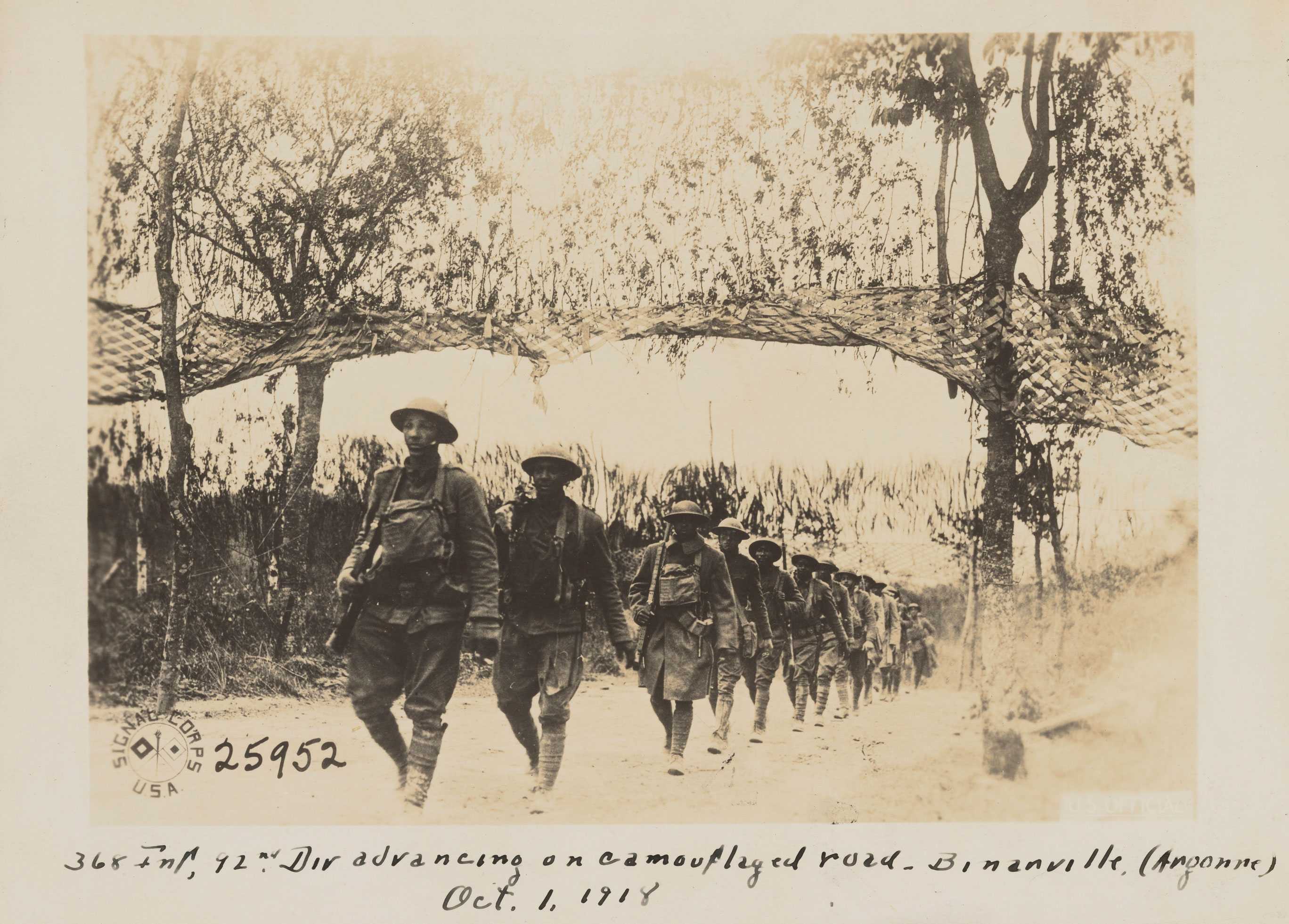 Soldiers walk single-file down a tree-flanked road, carrying rifles. Nets cover the road.