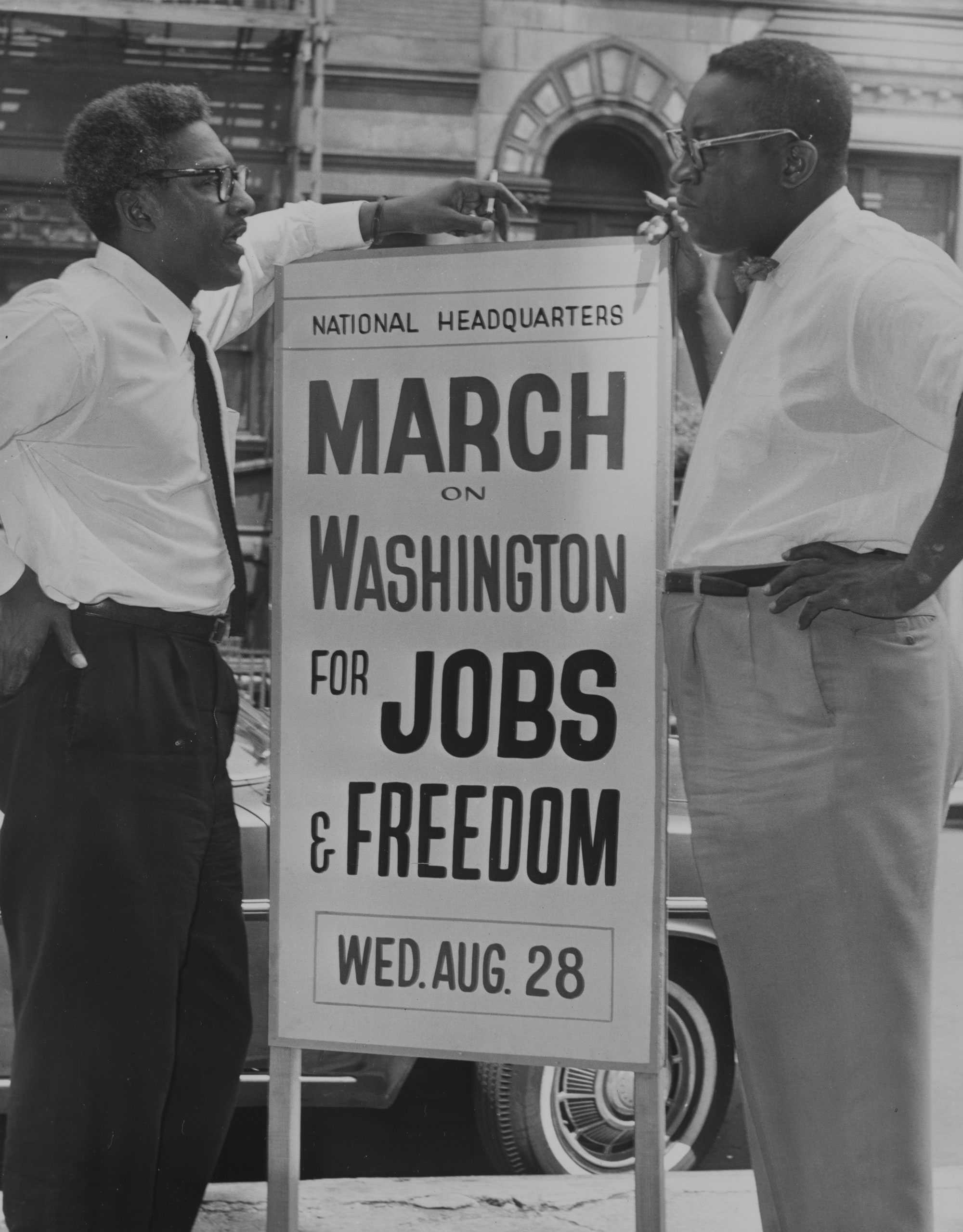 Bayard Rustin and Cleveland Robinson take a smoke break and rest their arms on a protest sign for the March on Washington for Jobs and Freedom as they talk together.