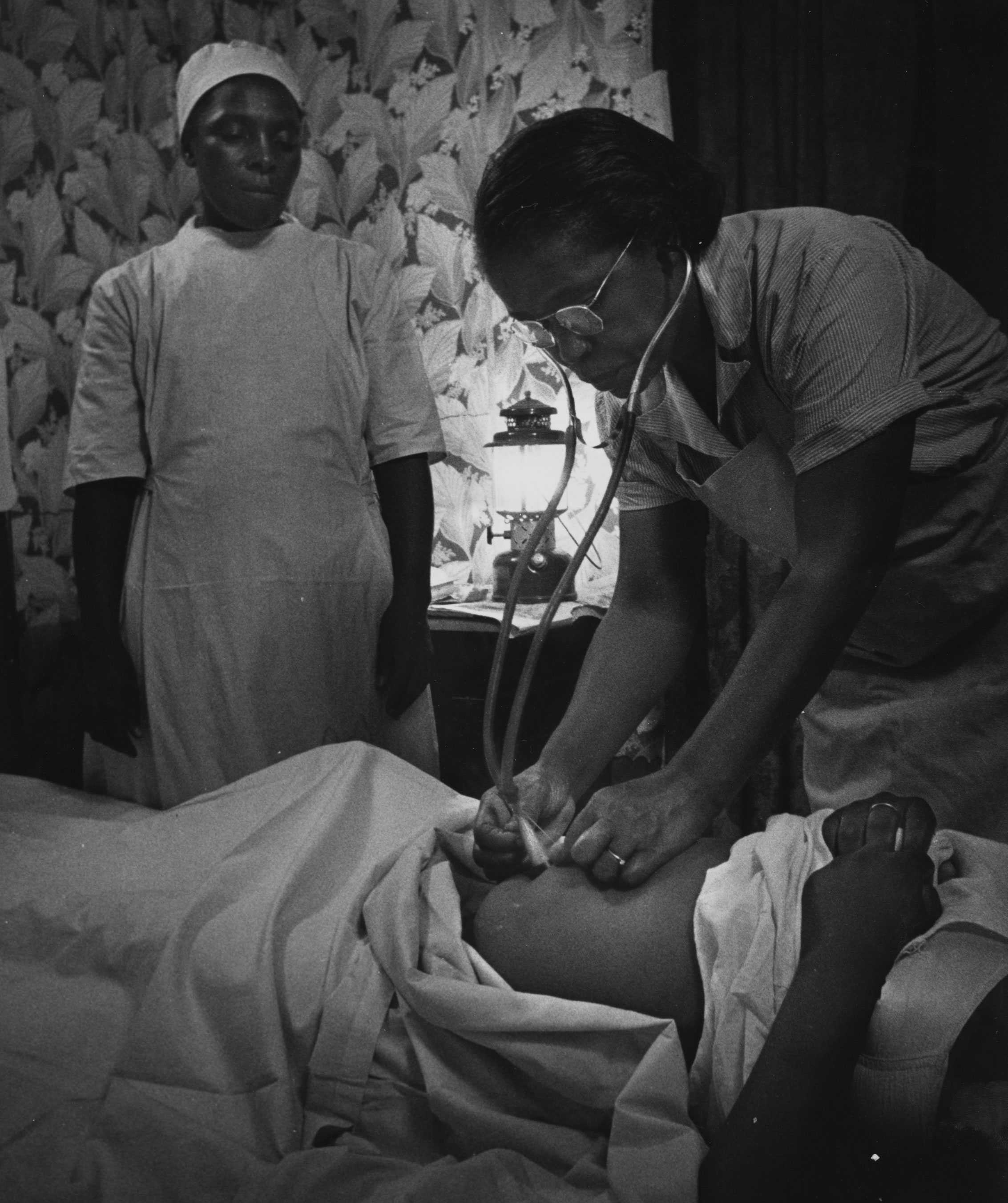 A black-and-white photograph of Maude Callen using a stethoscope on a patient's stomach. Another nurse is standing and watching.