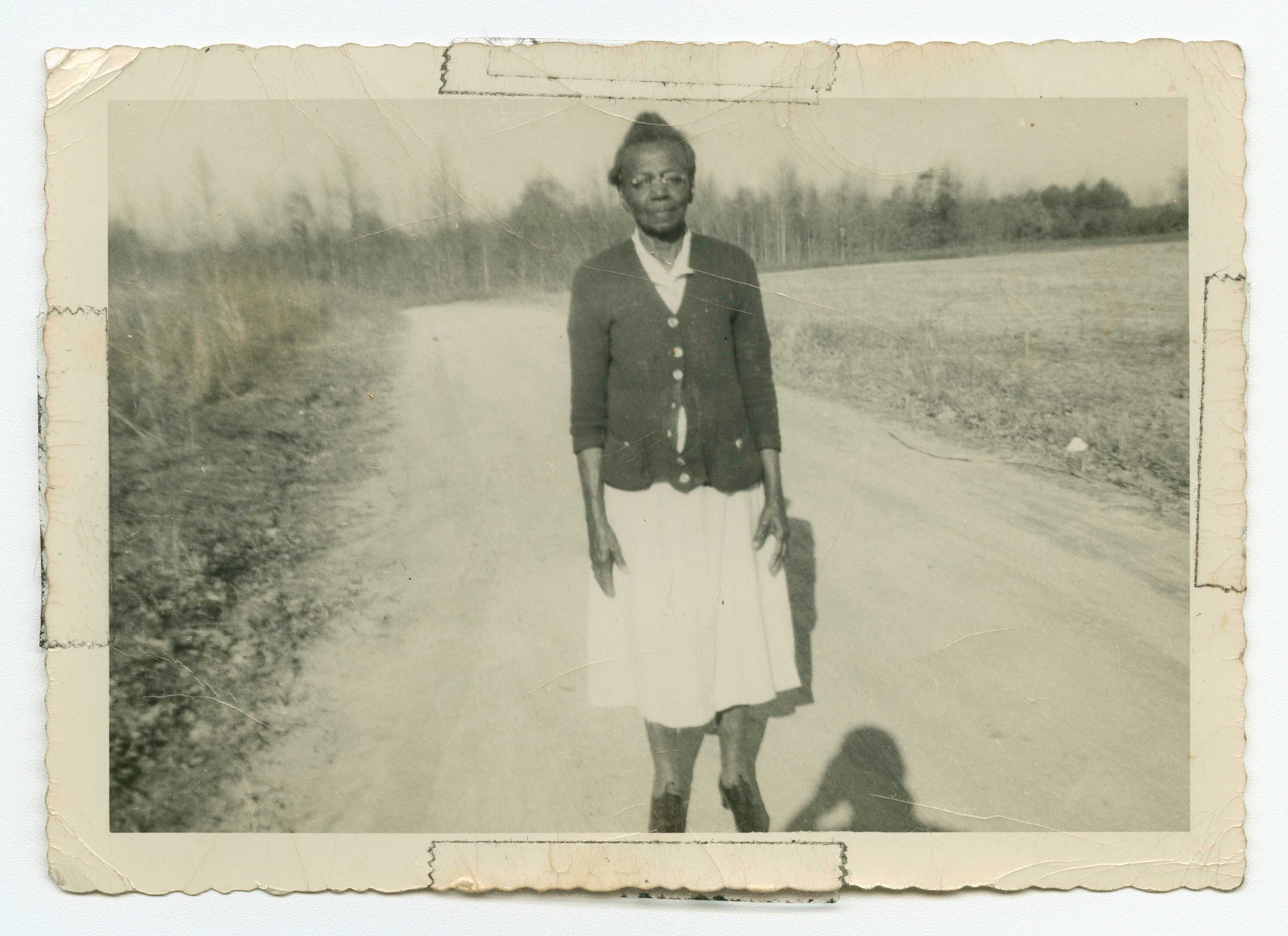 A black-and-white photograph of third-generation midwife Susie Carey, standing on a dirt road in a rural landscape.