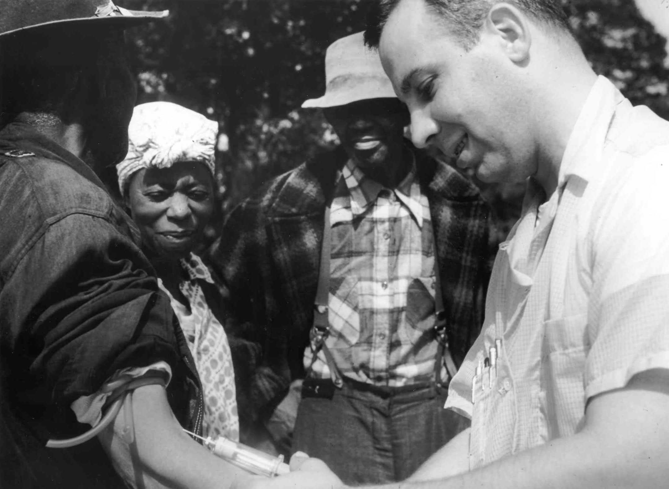 Black and white photograph showing an unidentified man having his blood drawn by a white man.  A woman and man look on.