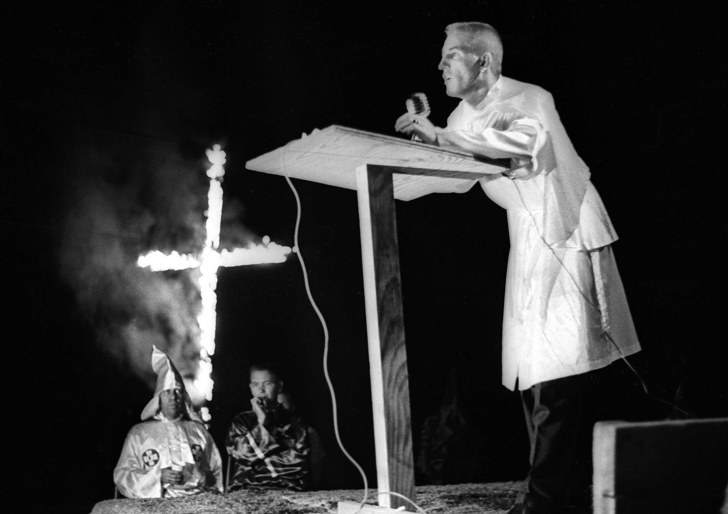 A black and white photograph of a KKK member speaking to a crowd as a large cross burns in the background.