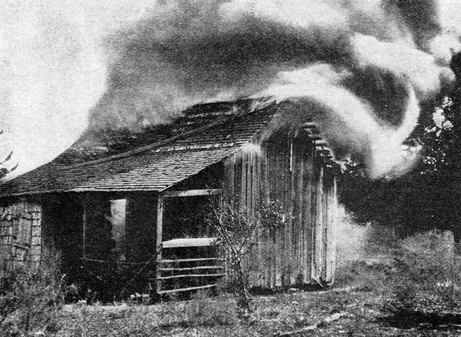 Black and white photograph showing small wooden home in flames.