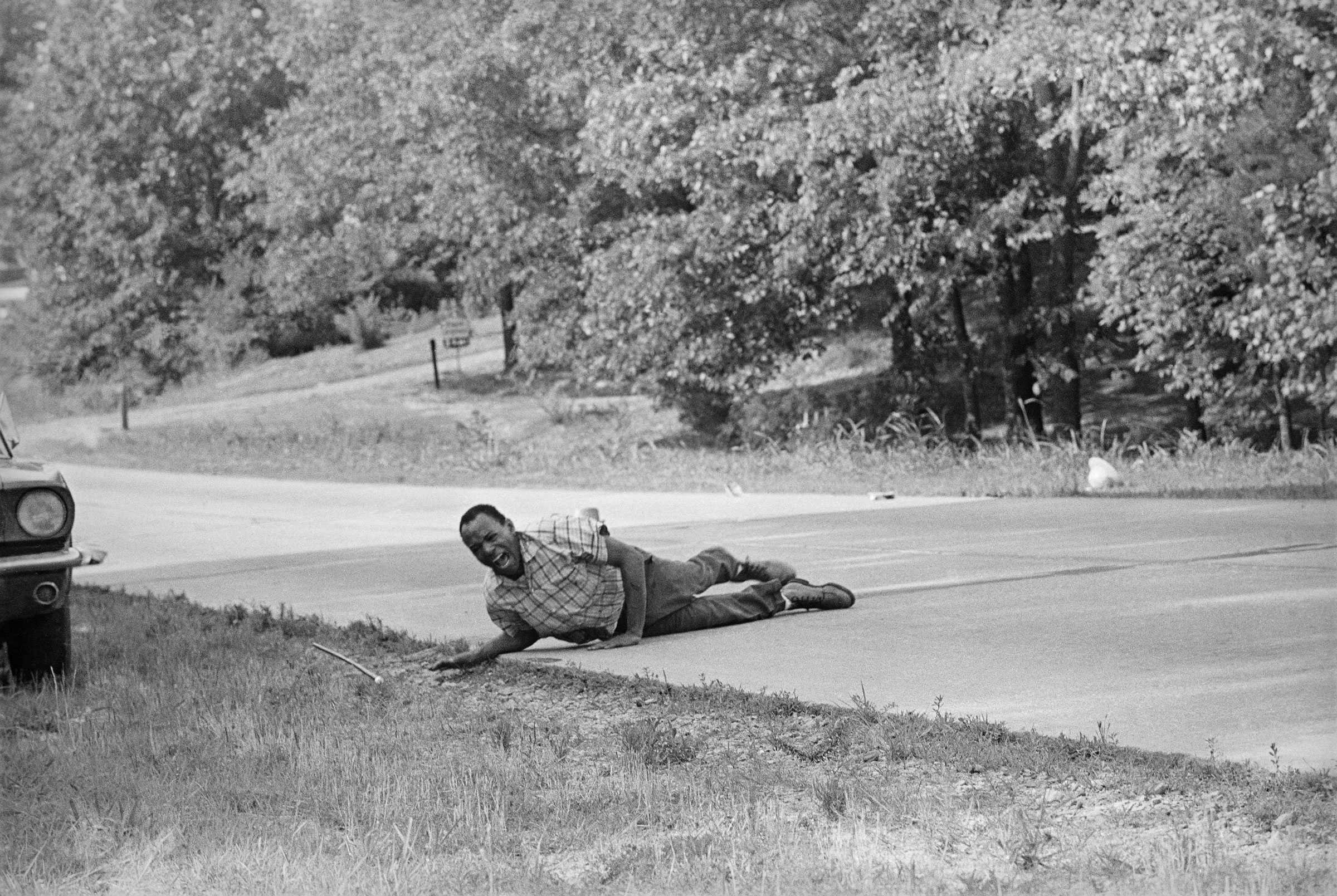 A black and white photograph of James Meredith grimaces in pain as he pulls himself across a Highway after being shot.