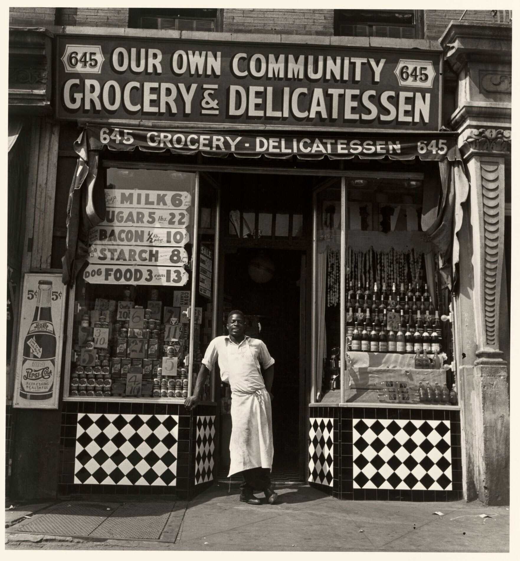 Black and white gelatin silver photographic print of a man wearing apron shown standing in doorway of storefront of "Our Own Community Grocery & Delicatessen".